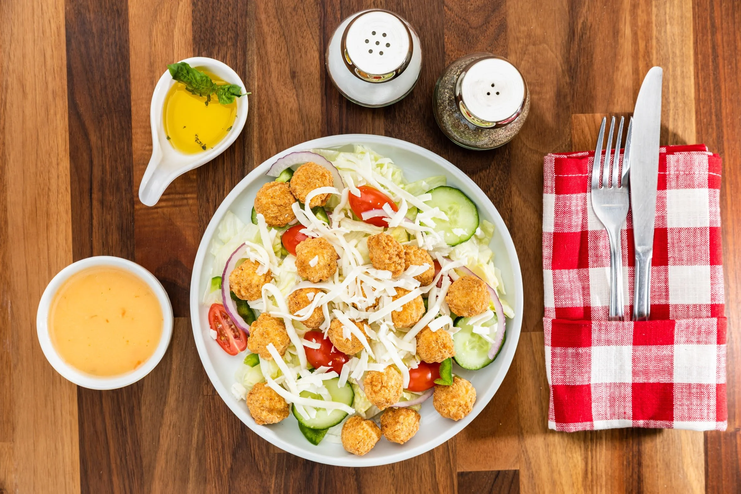 A fresh salad with shredded cheese, cherry tomatoes, cucumber slices, red onion, lettuce, and breaded chicken pieces in a white bowl on a wooden table. Accompanying the salad are a small bowl of salad dressing, a small dish of olive oil and balsamic 