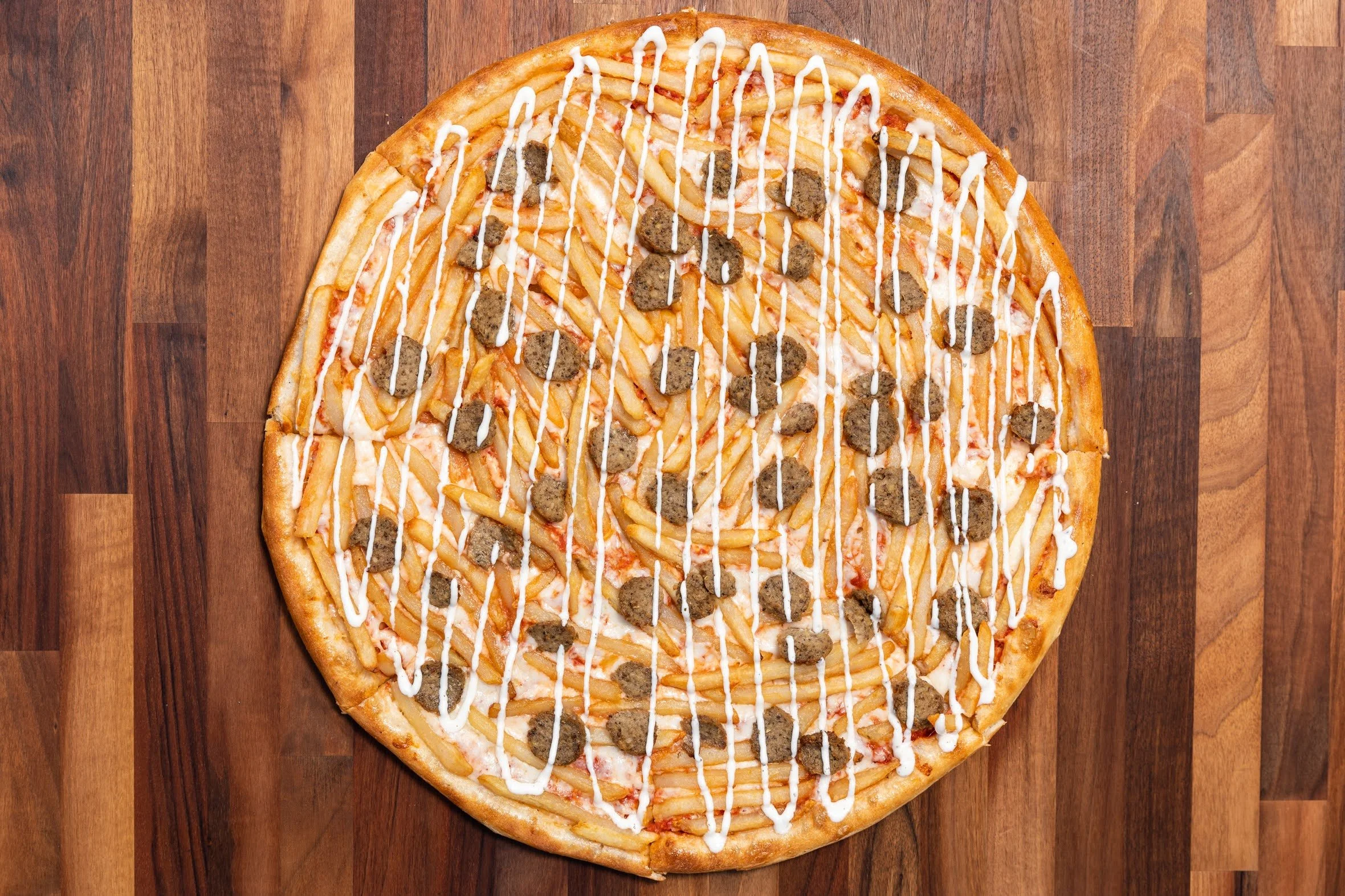 Pizza with French fries, sausage chunks, white sauce drizzle, and other toppings on a wooden surface.