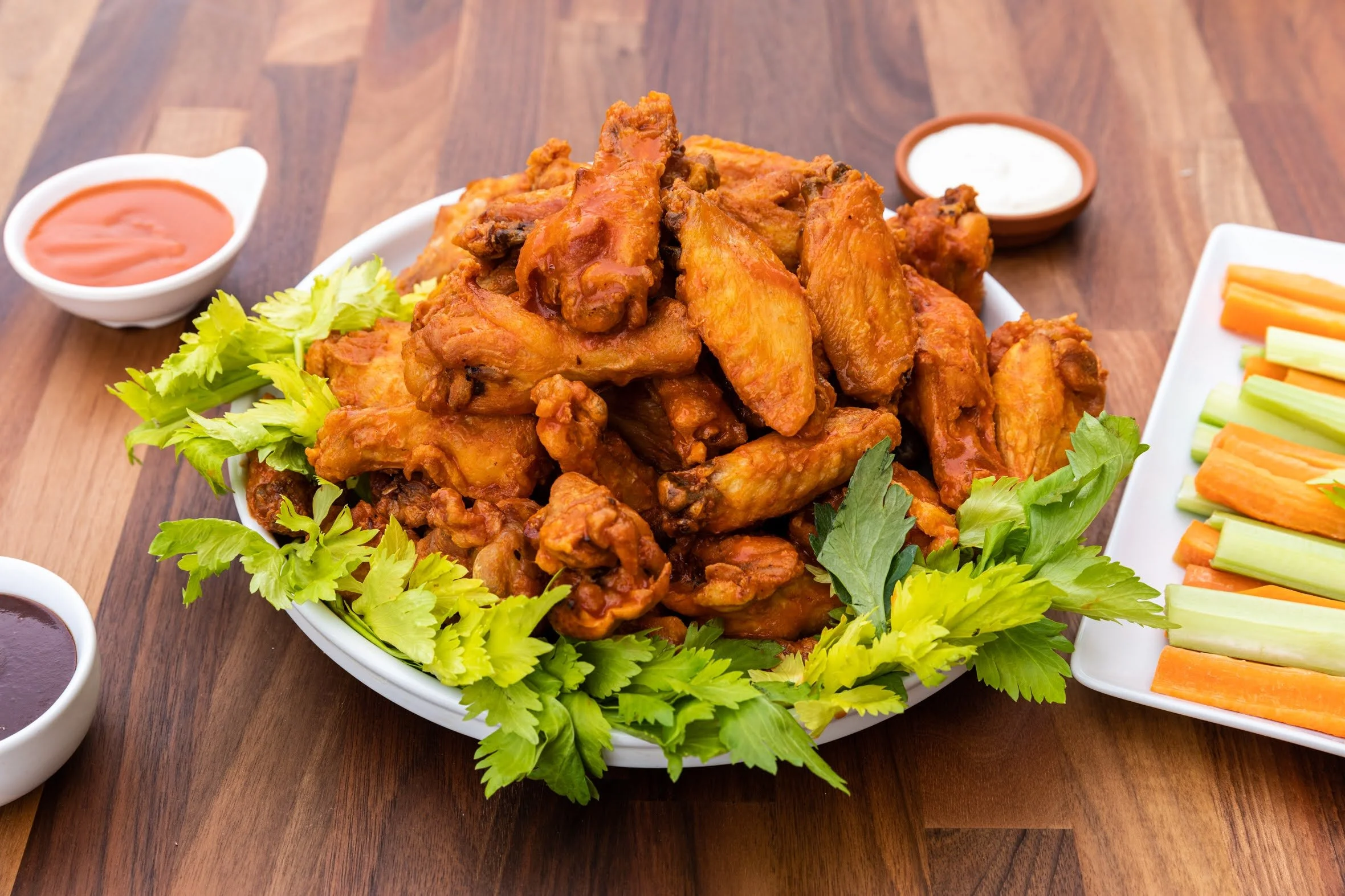 A plate of fried chicken wings garnished with fresh green herbs, with side dishes of dipping sauces and vegetable sticks on a wooden table.