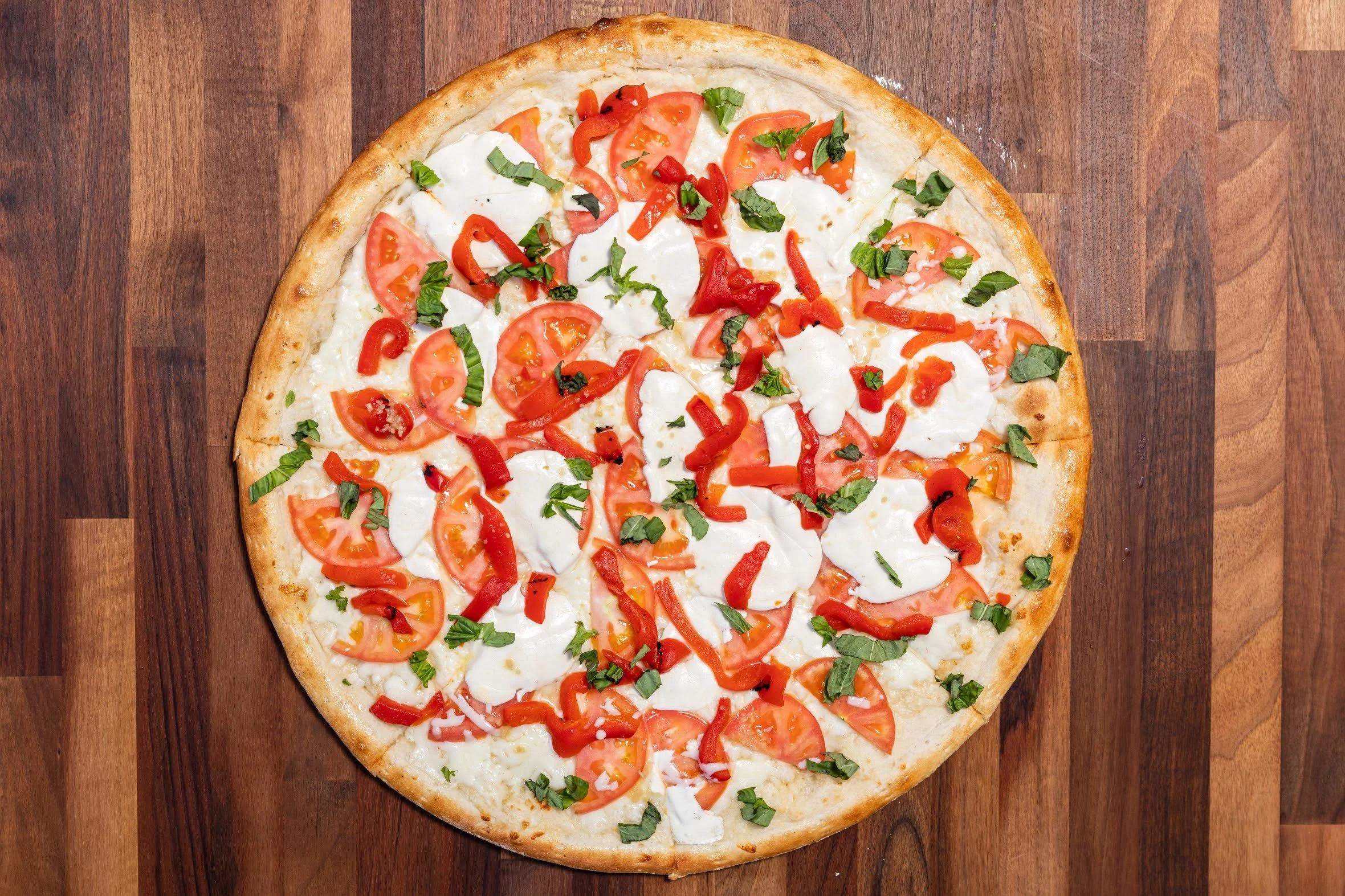 A pizza topped with tomato slices, red bell pepper strips, basil leaves, and dollops of mozzarella cheese on a wooden surface.