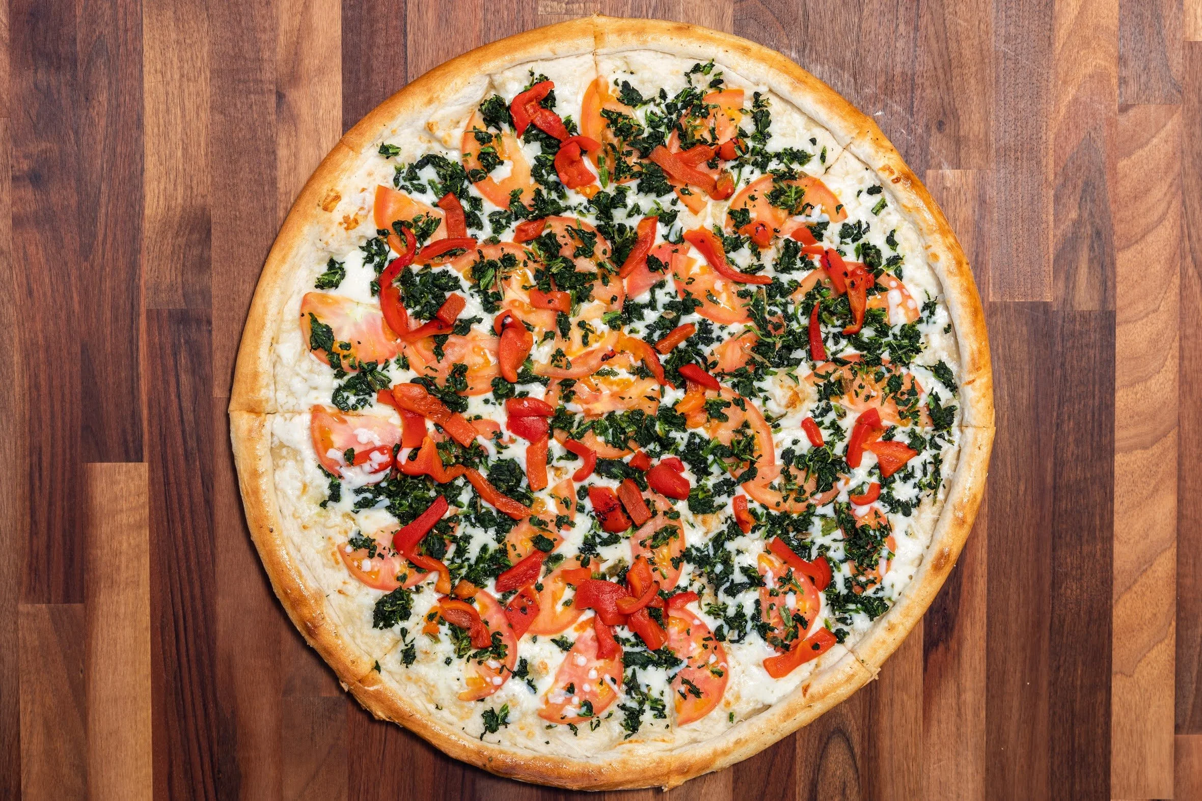 A pizza with toppings including sliced tomatoes, red bell peppers, and chopped kale on a wooden surface.