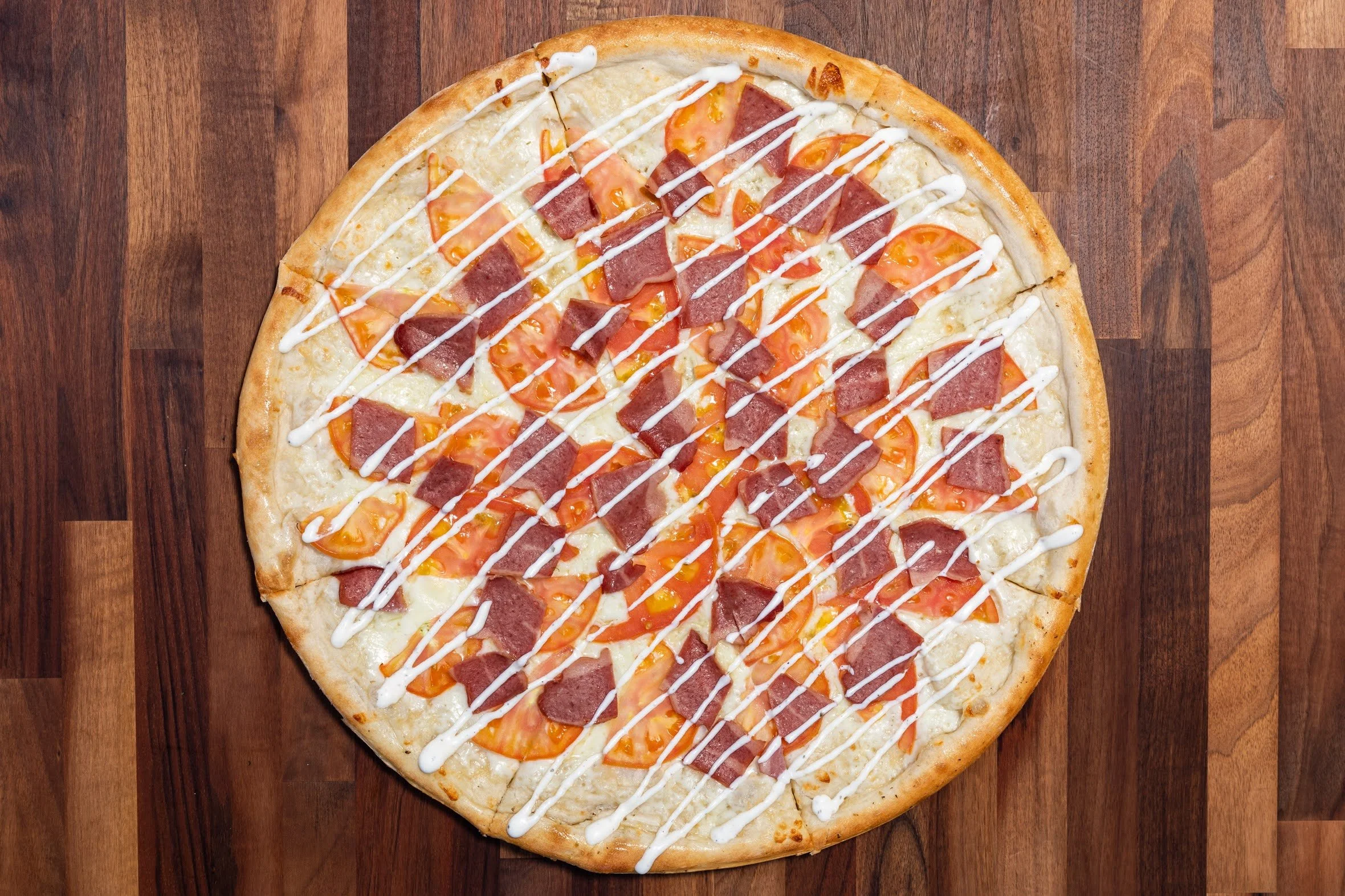 A pizza with tomato slices, ham, melted cheese, and drizzled white sauce on a wooden surface.