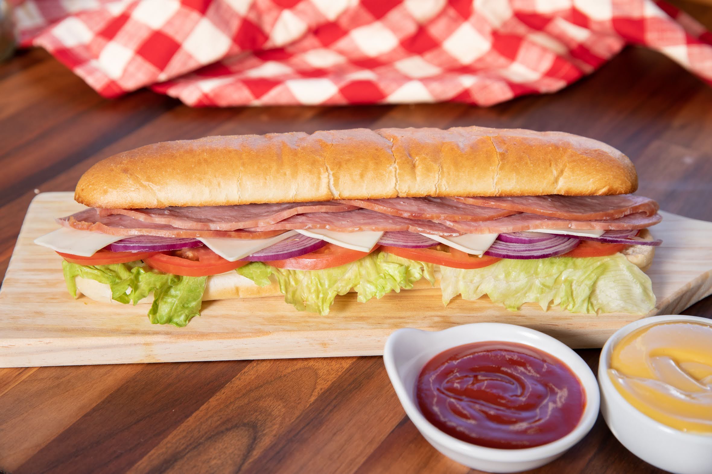 A large sub sandwich on a wooden board with lettuce, tomato, onions, cheese, ham, and bread, accompanied by bowls of ketchup and mustard on the side.