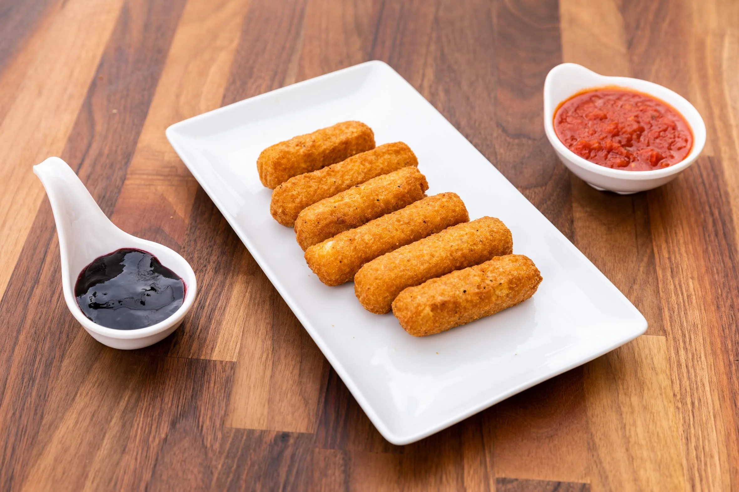 Six mozzarella sticks on a white rectangular plate with two dipping sauces, one red and one dark purple, on a wooden table.