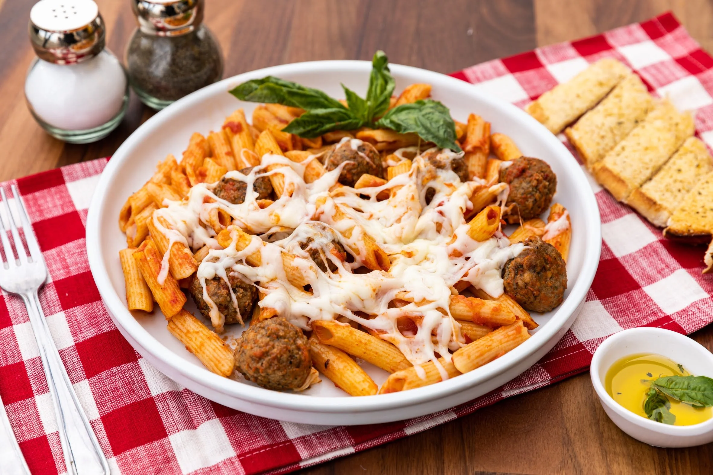 A plate of baked ziti pasta with meatballs and melted cheese, garnished with basil, served with slices of garlic bread, a small bowl of olive oil with herbs, and salt and pepper shakers on a red and white checkered tablecloth.