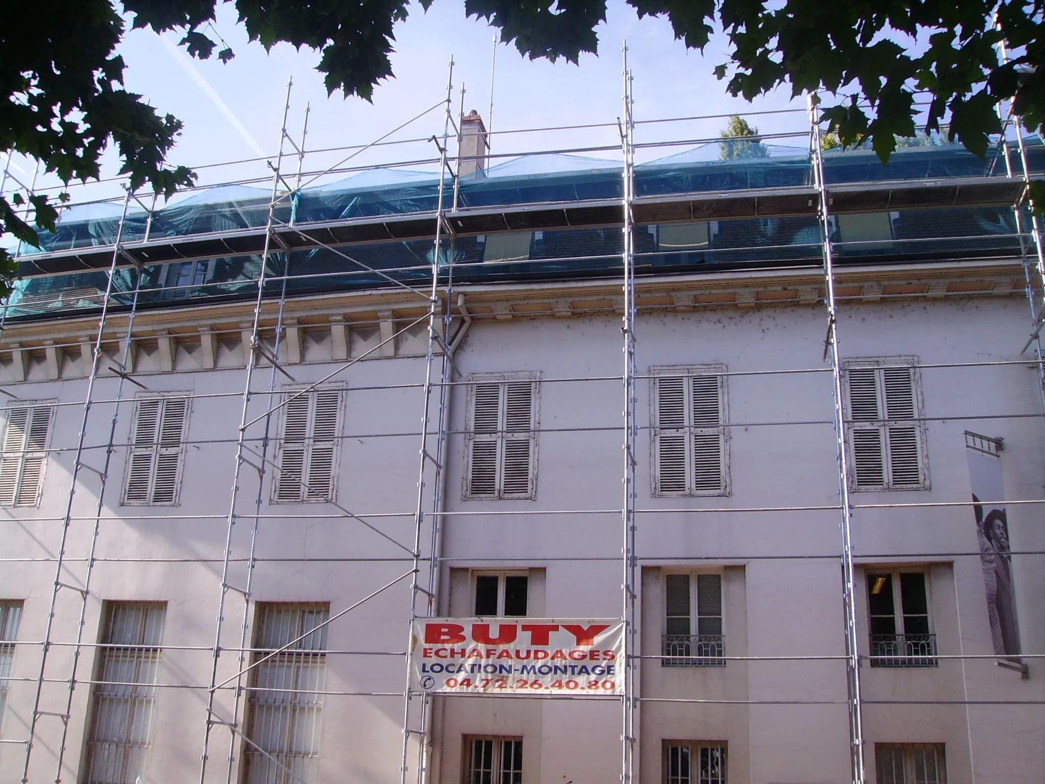 Une façade d'immeuble en travaux, entourée d'une structure d'échafaudages en métal. Il y a un panneau indiquant la location et la pose d'échafaudages, avec un numéro de téléphone.