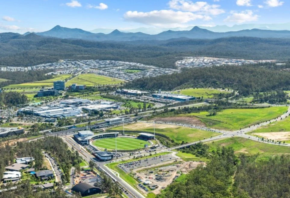 Aerial view of a sports stadium surrounded by roads, green fields, and surrounding buildings with mountains in the background under a cloudy sky.