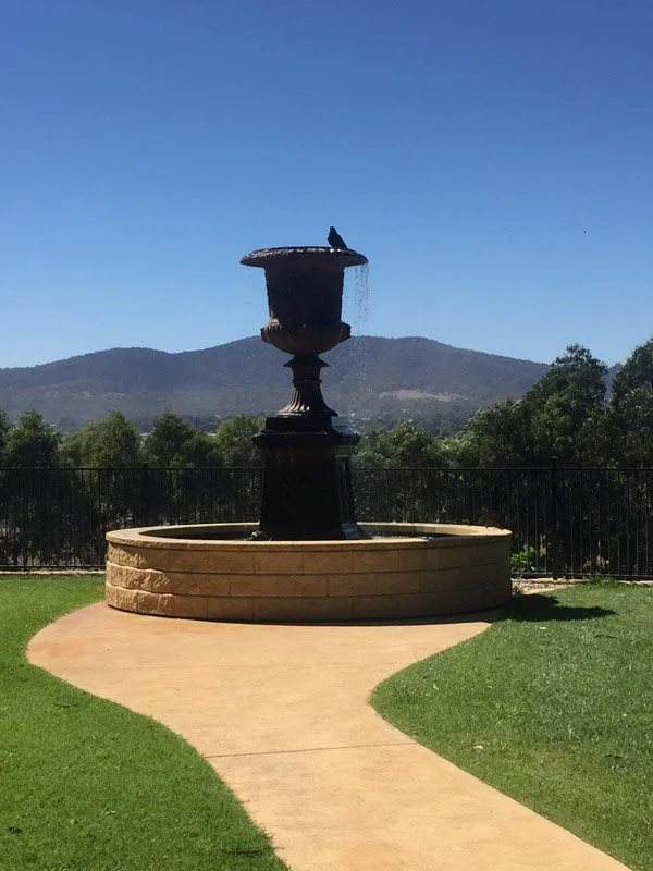 A black fountain with water flowing from the top, situated on a circular stone platform, with a bird perched on its upper edge, set in a grassy park area with a winding pathway leading up to it, and trees and mountains in the background under a clear blue sky.