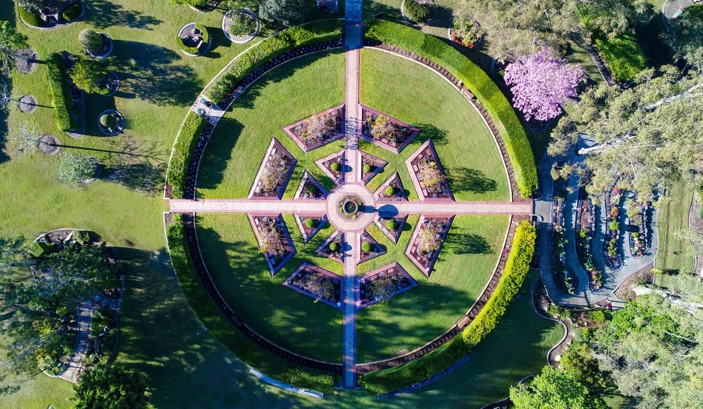 An aerial view of a circular park with a geometric flower-shaped centerpiece, surrounded by walkways, green lawns, trees, and parking lots.