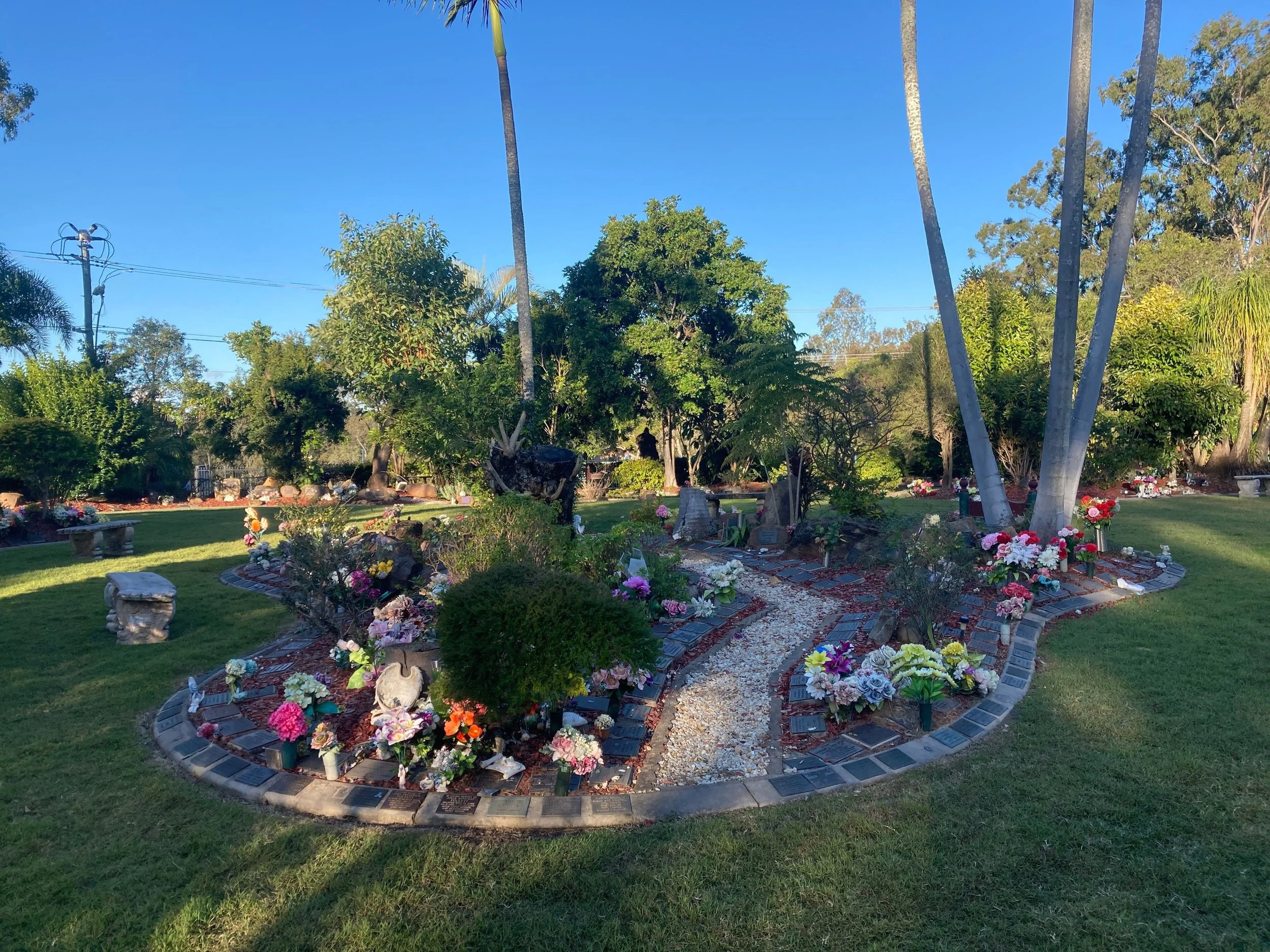 A cemetery with a circular arrangement of small headstones and flowers at sunset. Tall trees and a utility pole are visible in the background.