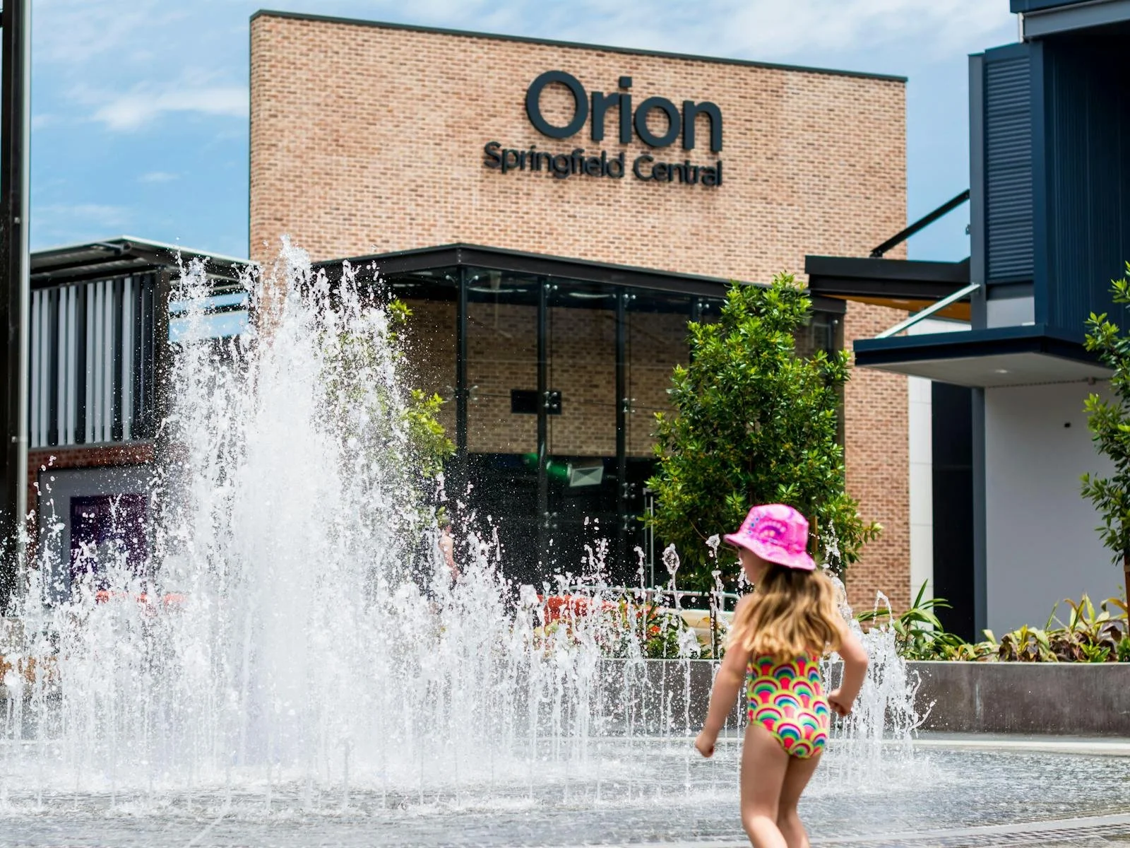 Child in a rainbow-colored swimsuit and pink sun hat playing in a water fountain outside Orion Springfield Central shopping mall.
