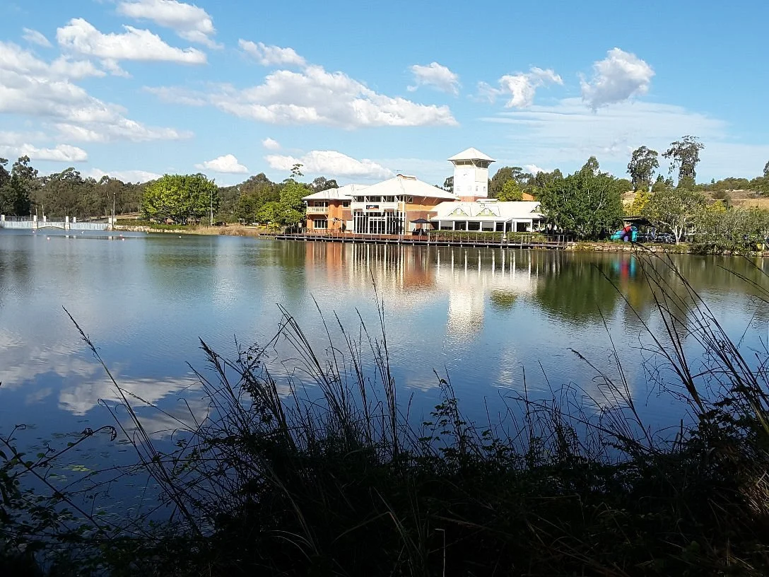 A lakeside building with a tower, surrounded by trees and reflected in the water, under a partly cloudy sky.
