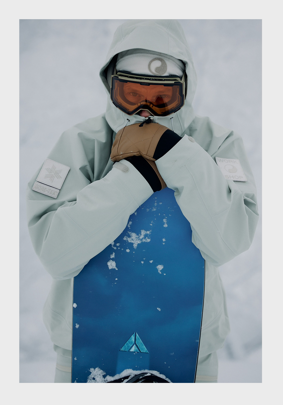 A person dressed in white winter gear, including a hood and goggles, holding a snowboard against a snowy background