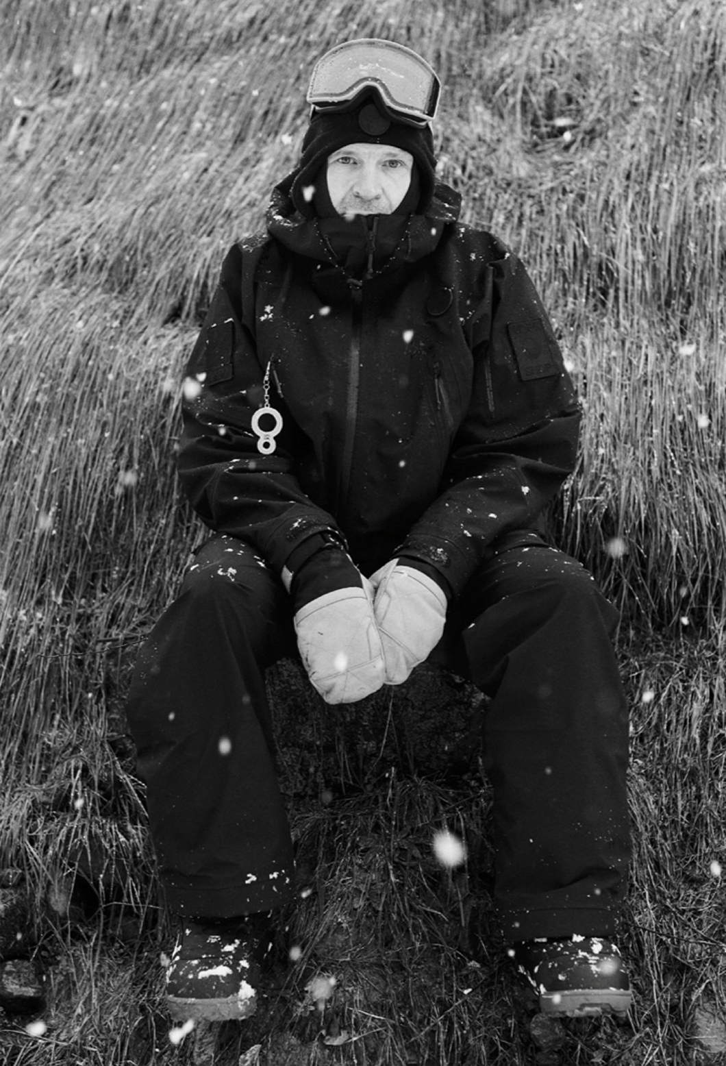 A person dressed in winter gear sits on the ground in a grassy, snowy landscape. They wear goggles on their head, a black jacket, gloves, and snow boots, with snow falling around them.