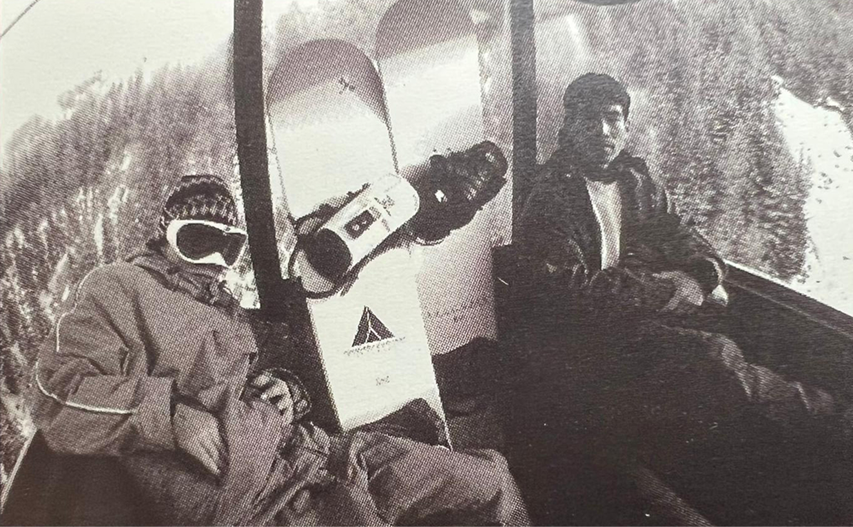 Black and white photo of two people sitting on a bench outdoors. One person is wearing a jacket with goggles or glasses, the other person is dressed in a dark jacket and light shirt. Between them is a small table with beverages and a paper bag.