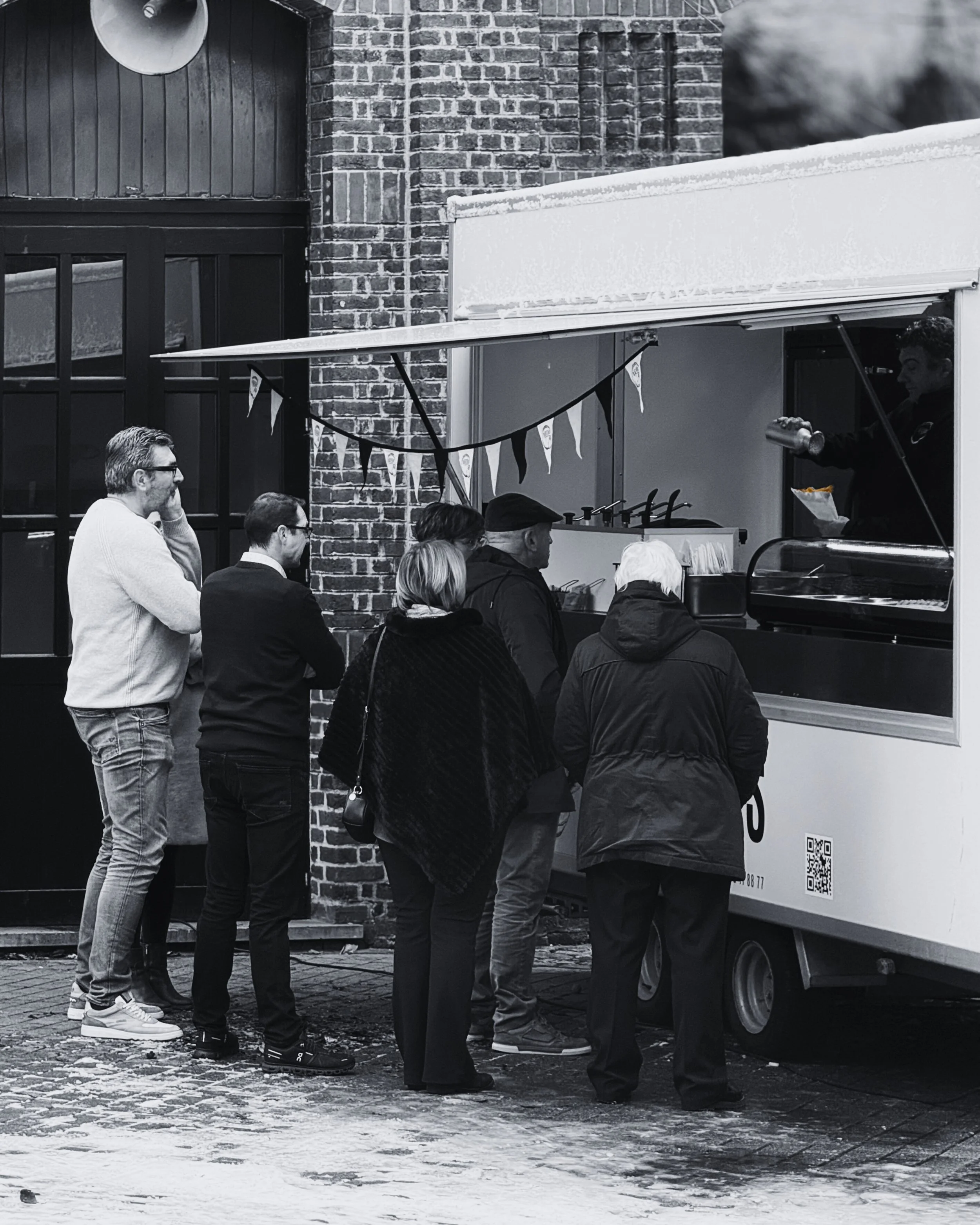 Mensen staan in een rij voor een foodtruck, die eten serveert. Het is een zwart-wit foto.