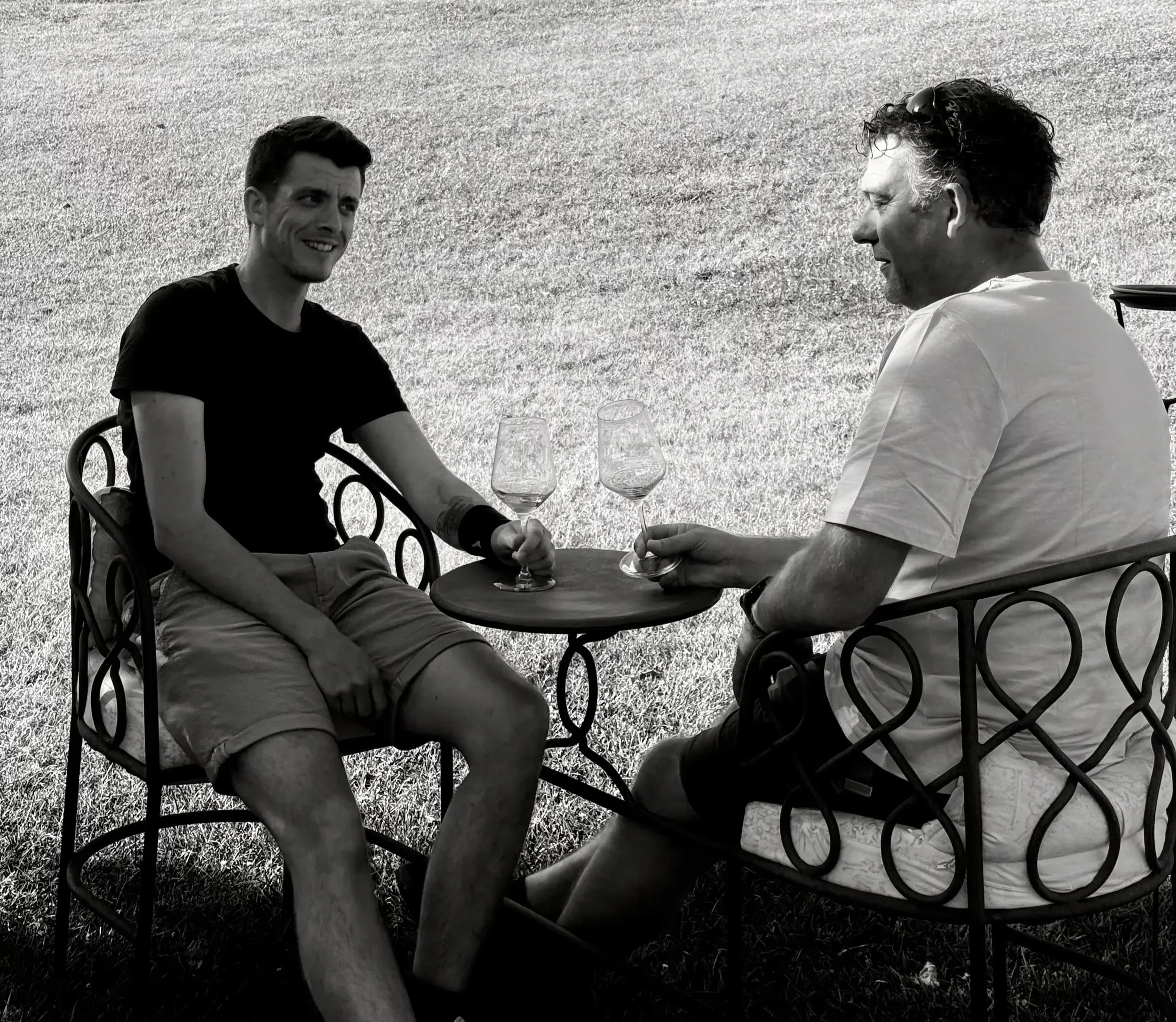 Twee mannen zitten buiten op een terras met metalen stoelen, praten en drinken uit wijnglazen.