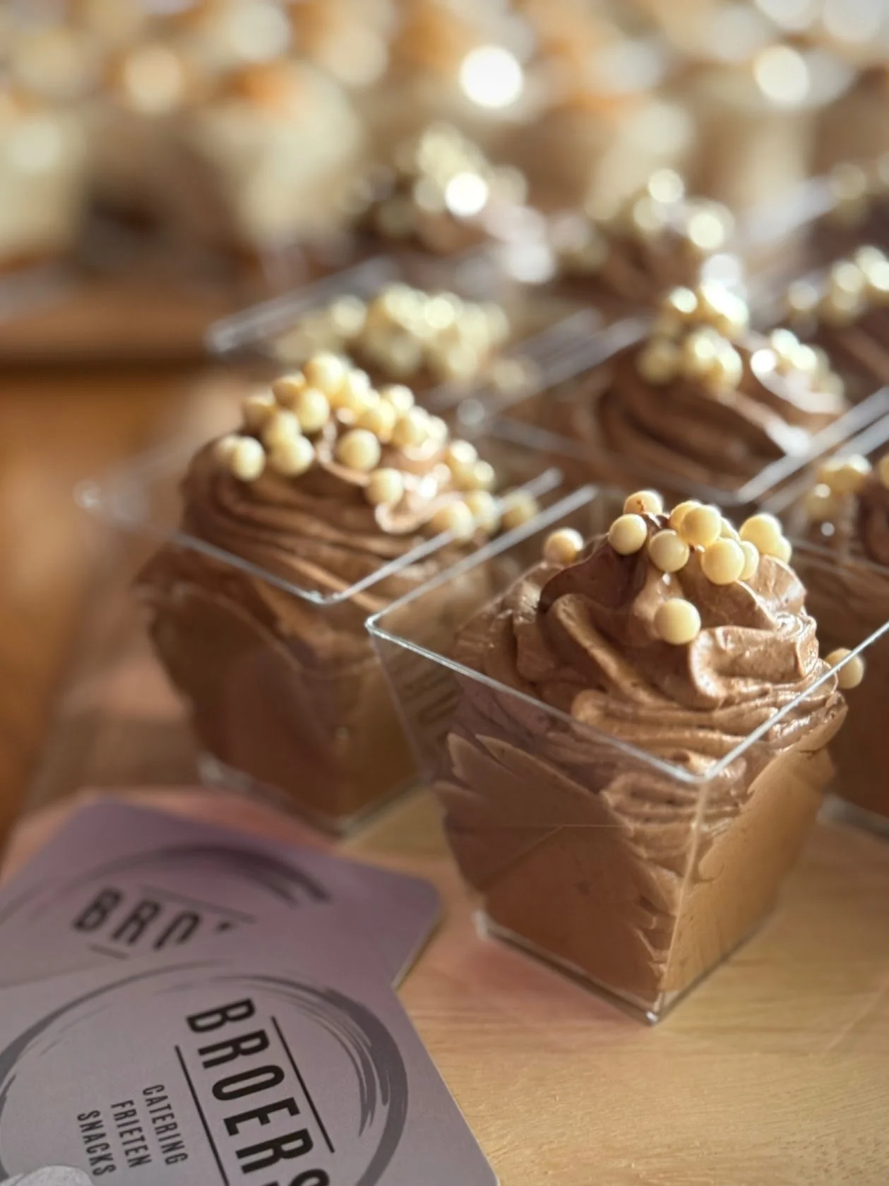 Chocolademousse in kleine vierkante doorzichtige bekers met witte parels bovenop.