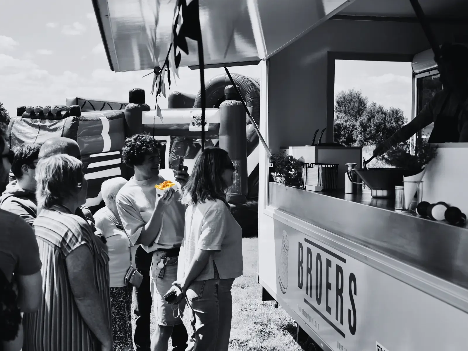 In een zwart-wit foto staan mensen in een rij buiten bij een foodtruck met het woord 'BROERS' op de zijkant; een persoon serveert eten.