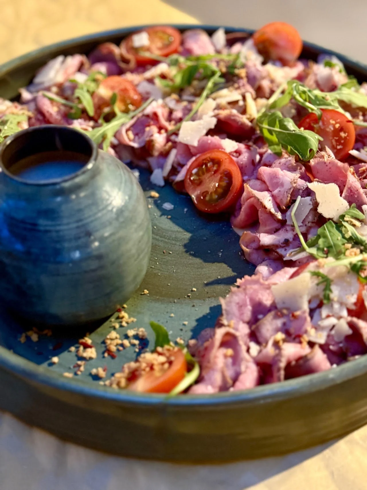 Salade met cherrytomaten, rucola, parmezaanse kaas en plakjes rauwe ham op een blauw bord, met een klein groene vaas of beker erbij.