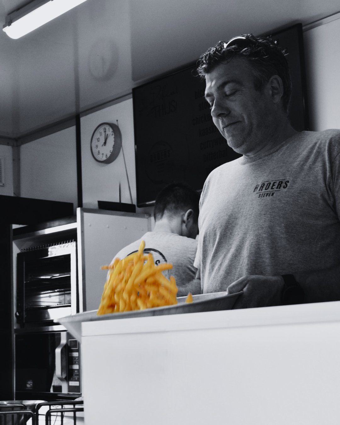 Man in een keukenkastje met een bak met gefrituurde frietjes, op de achtergrond een analoge klok en een tv-scherm.