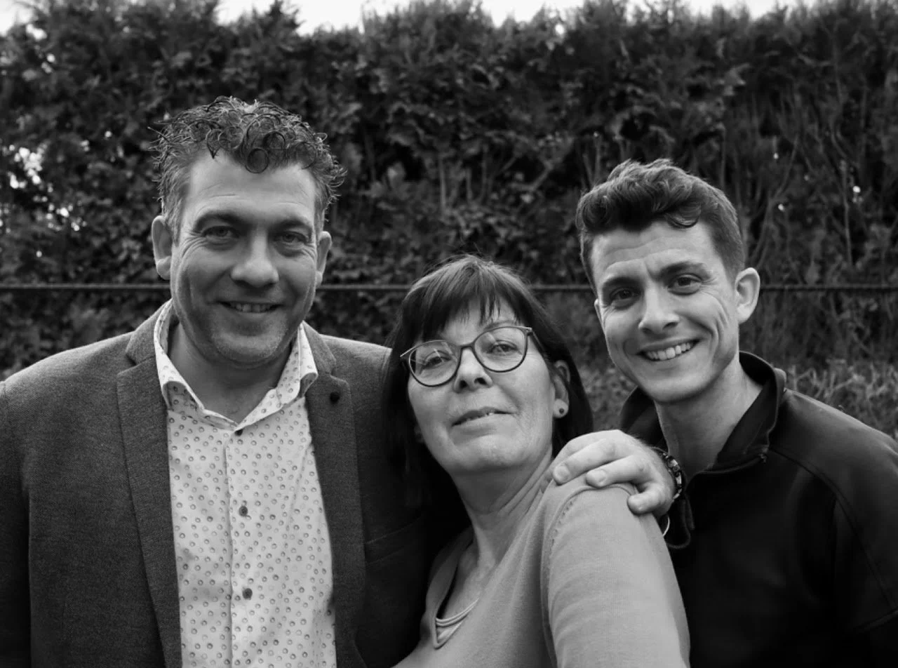 Driemensen vrouwen en mannen die samen naar de camera kijken en lachen, buiten met een donkergroen struik op de achtergrond.