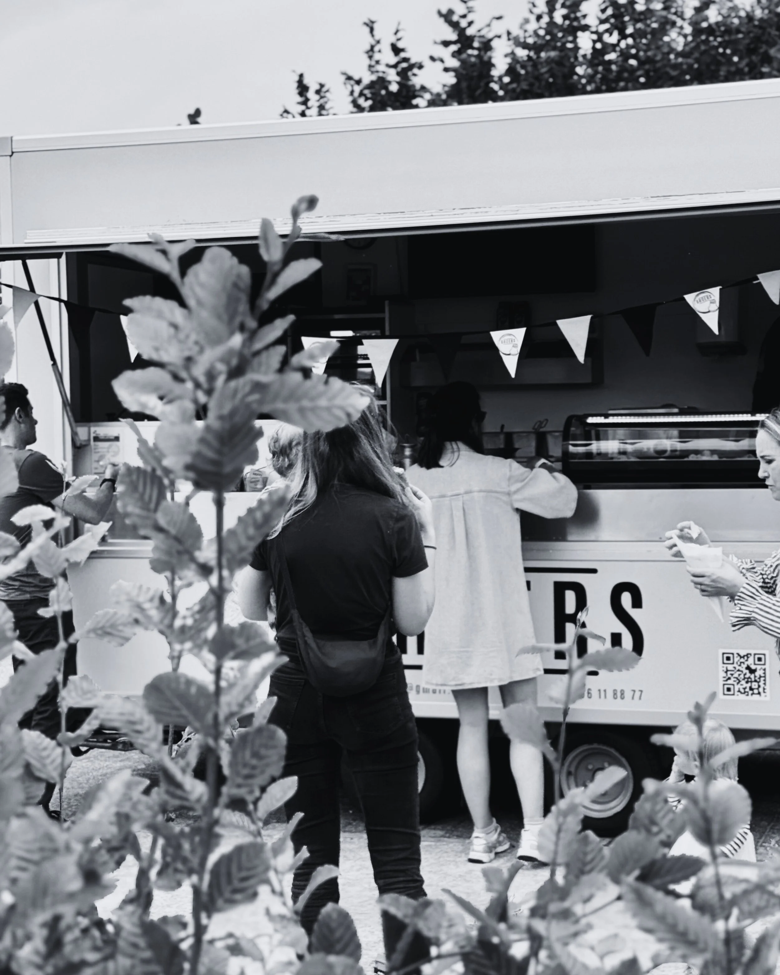 Mensen staan in rij bij een foodtruck, gezien door bladeren op de voorgrond, zwart-wit foto.