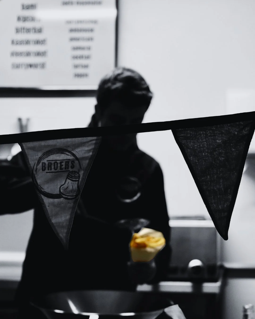 Persoon in een keuken, donker silhouet, houdt een geel bord vast, voor een zwart en wit achtergrond met een menu en vlaggetjes met het woord 'Broers'.