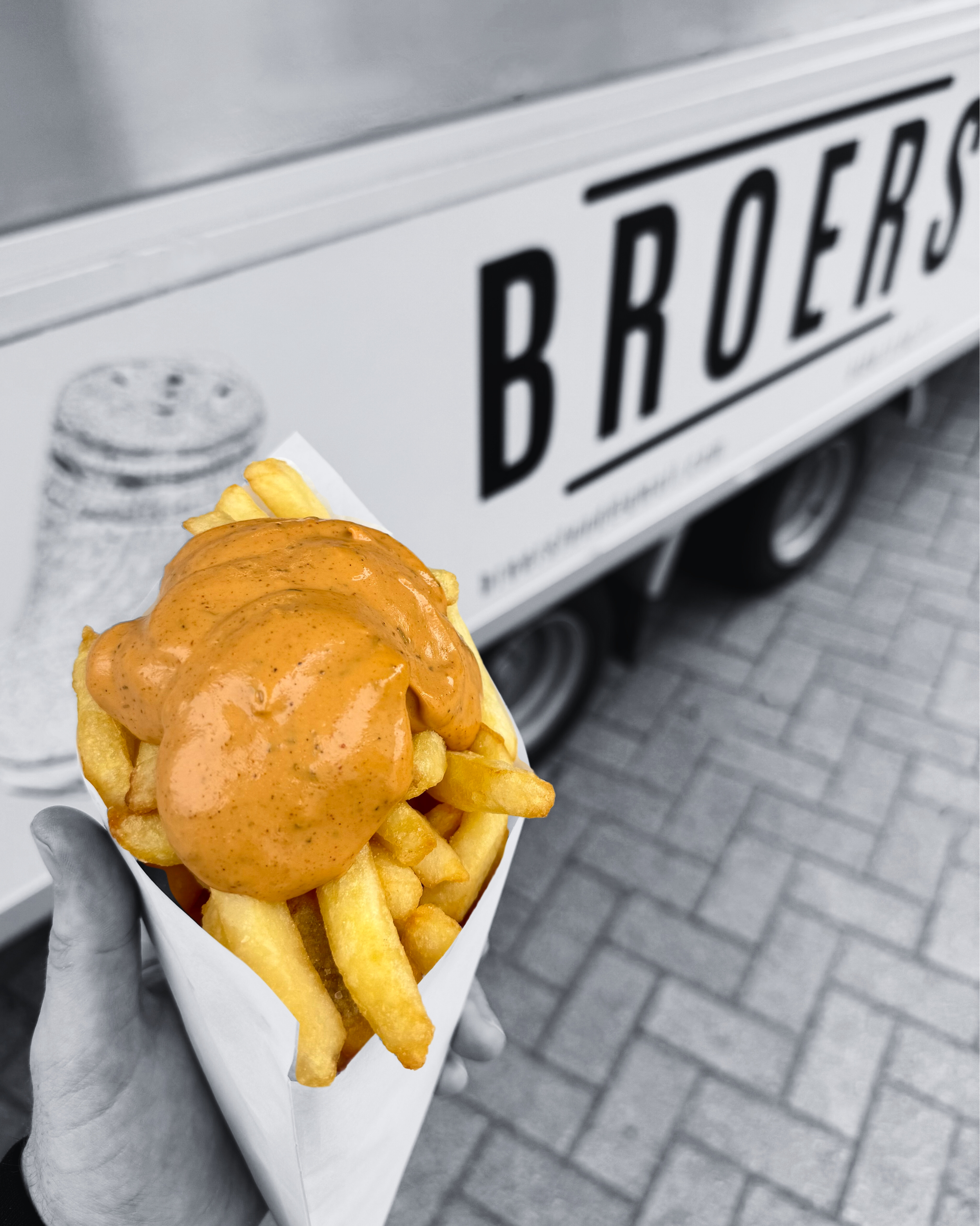 Een hand houdt een portie friet met een erbij gespoten saus voor een foodtruck met de naam 'B R O E R S' in de achtergrond