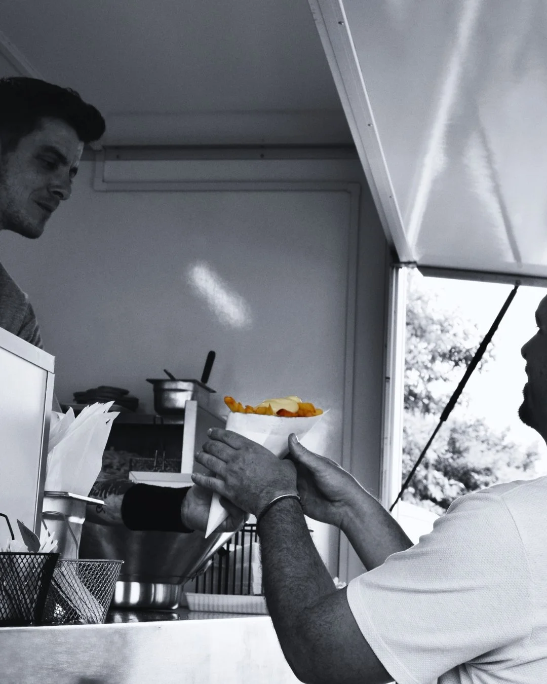 Man krijgt een schaal met hotdog van een ander man in een food truck of stand, zwart-wit foto met enkele kleurige hotdog toppings.