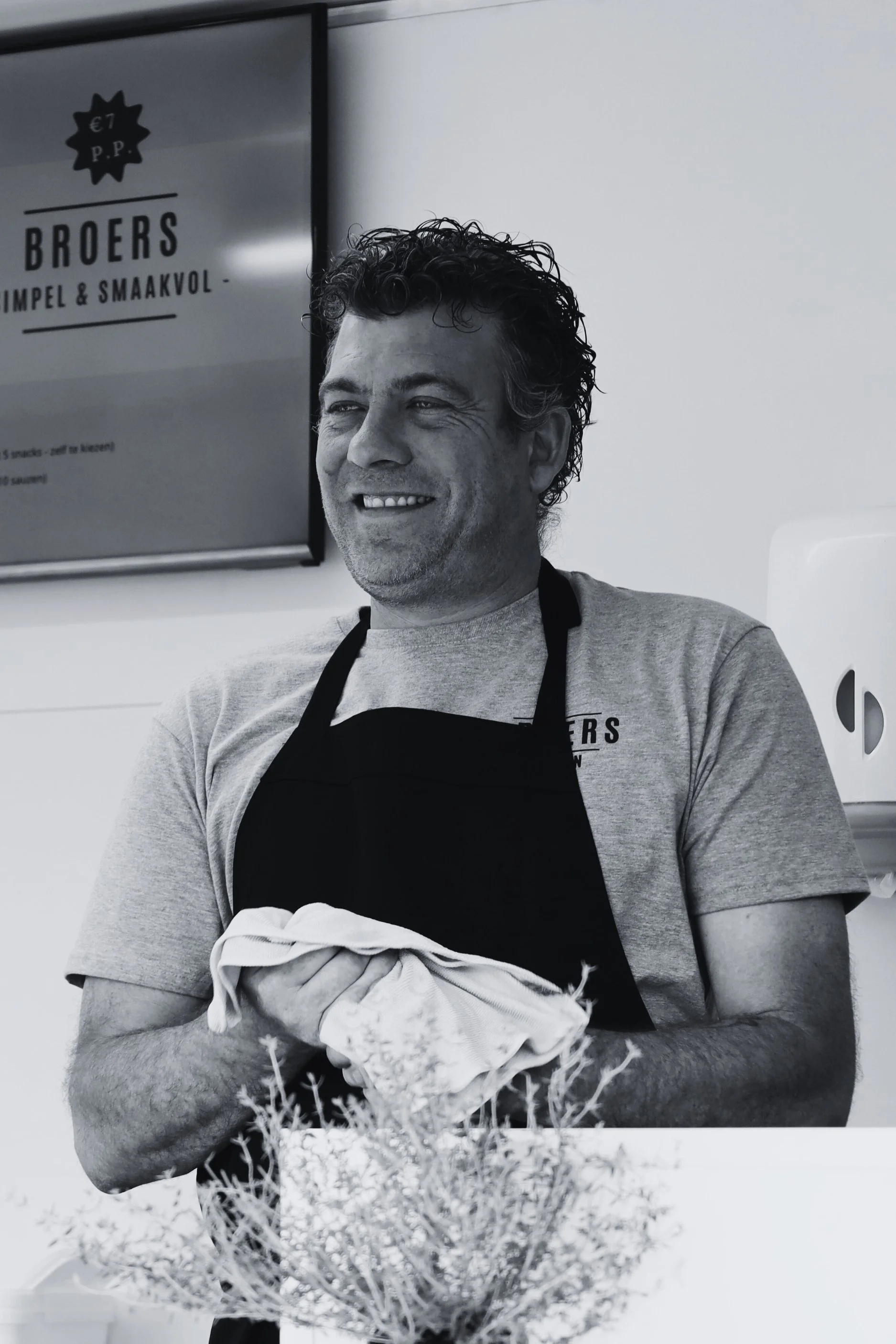 Man met krullend haar, glimlachend, draagt een grijs T-shirt en een zwarte schort, houdt een doek in zijn handen, staat in een keuken of winkel met een bord op de muur waarop 'Broers' staat.