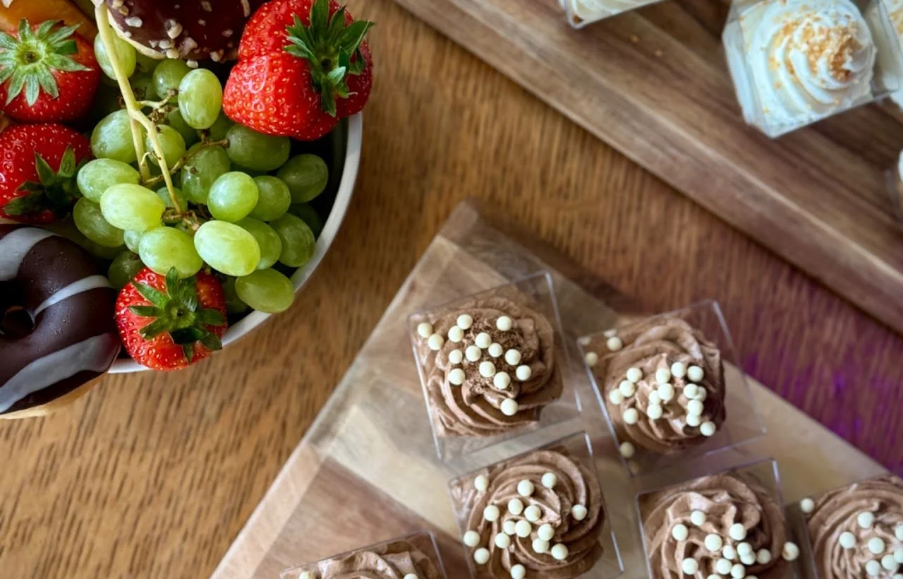 Een schaaltje met aardbeien en groene druiven, naast Kleine chocolade cupcakes versierd met witte balletjes op een houten tafel.
