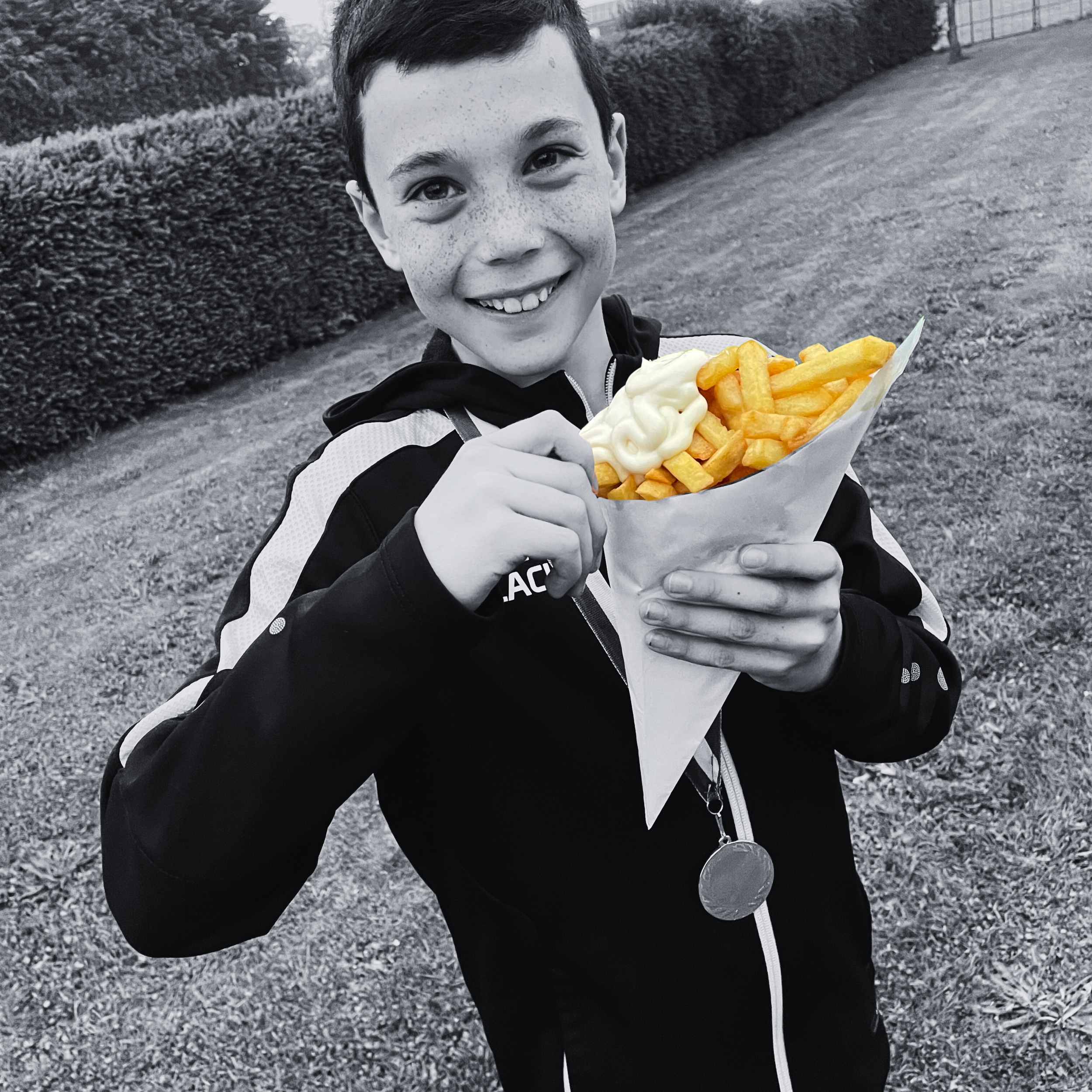 Een lachende jongen die ketchup en mayo saus op patat houdt, in een park met gras en heg op de achtergrond.