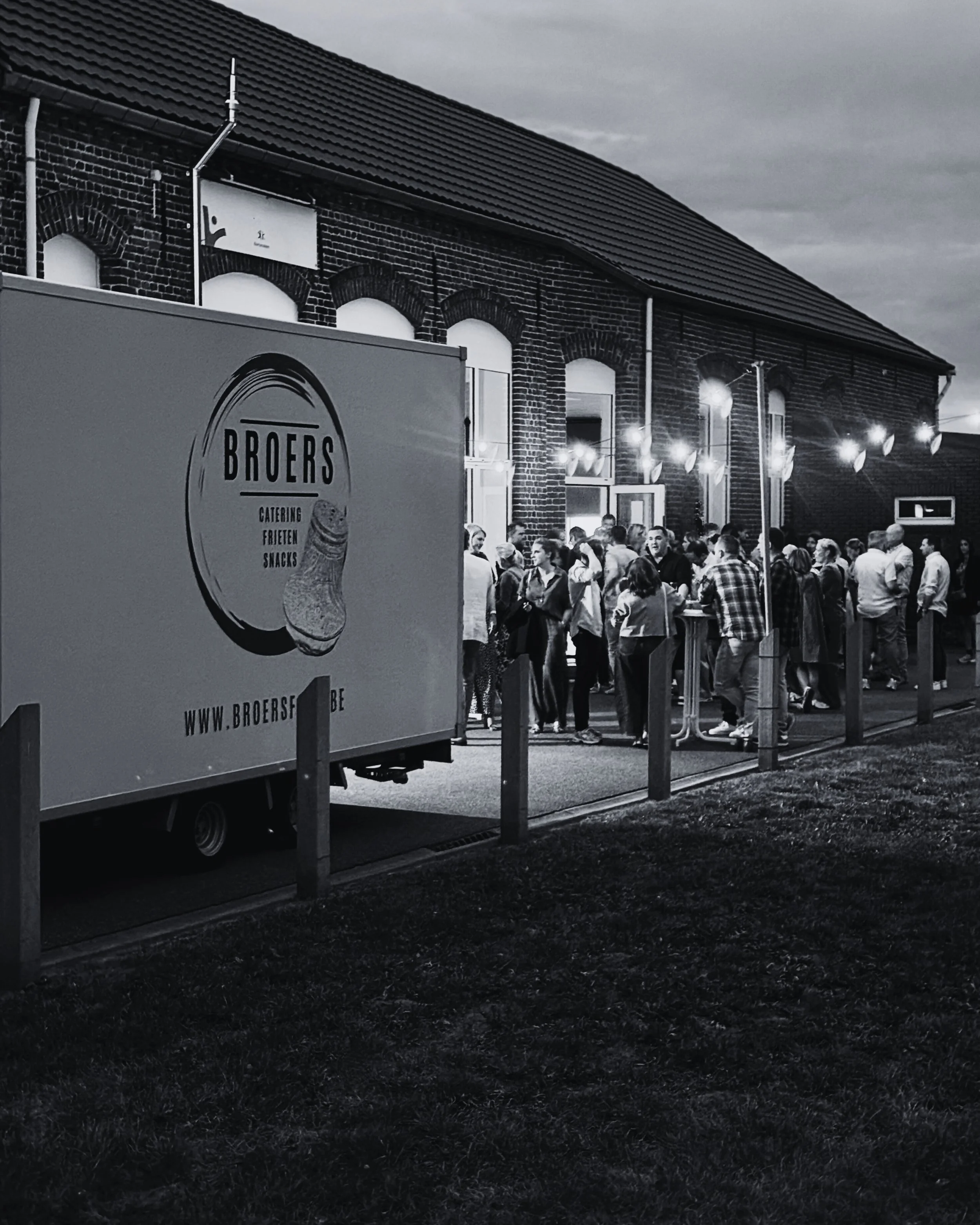 Grote groep mensen staat in een rij voor een gebouw, mogelijk wachtend op binnenkomst, met een vrachtwagen op de voorgrond die de branding 'Broers' heeft. Buitenverlichting hangt boven de menigte.