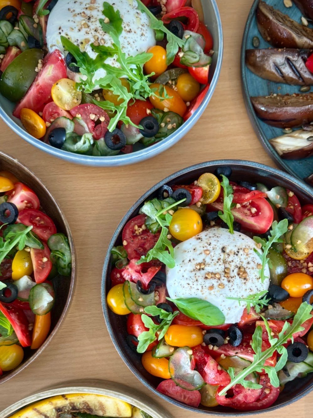 Salades met cherry tomaten, rucola, olijven en een gepocheerd ei, geserveerd in zwarte en blauwe schalen op een houten tafel.