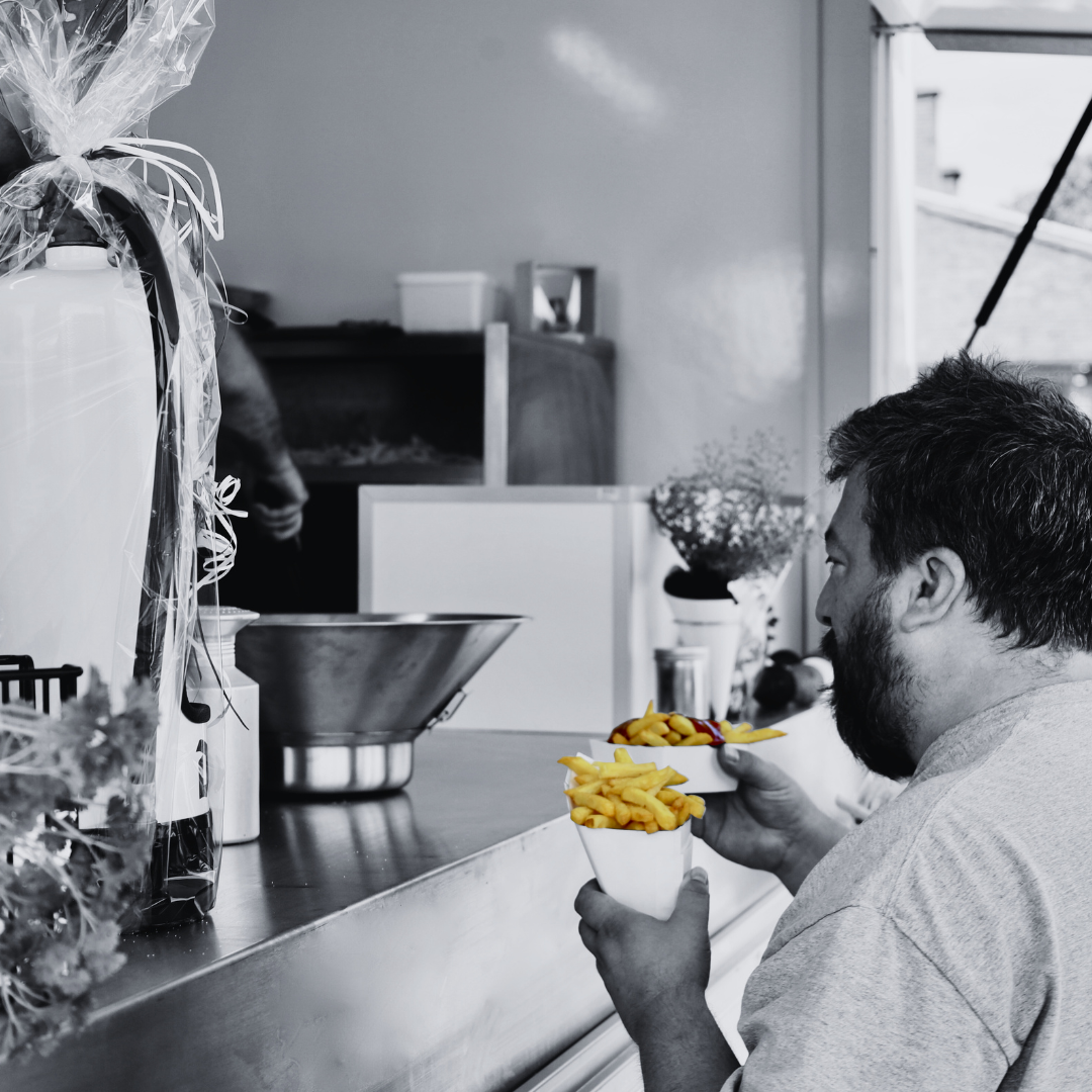 Man die frietjes eet uit een kartonnen beker, in een keuken of eetruimte met diverse objecten op het aanrecht en een raam op de achtergrond.