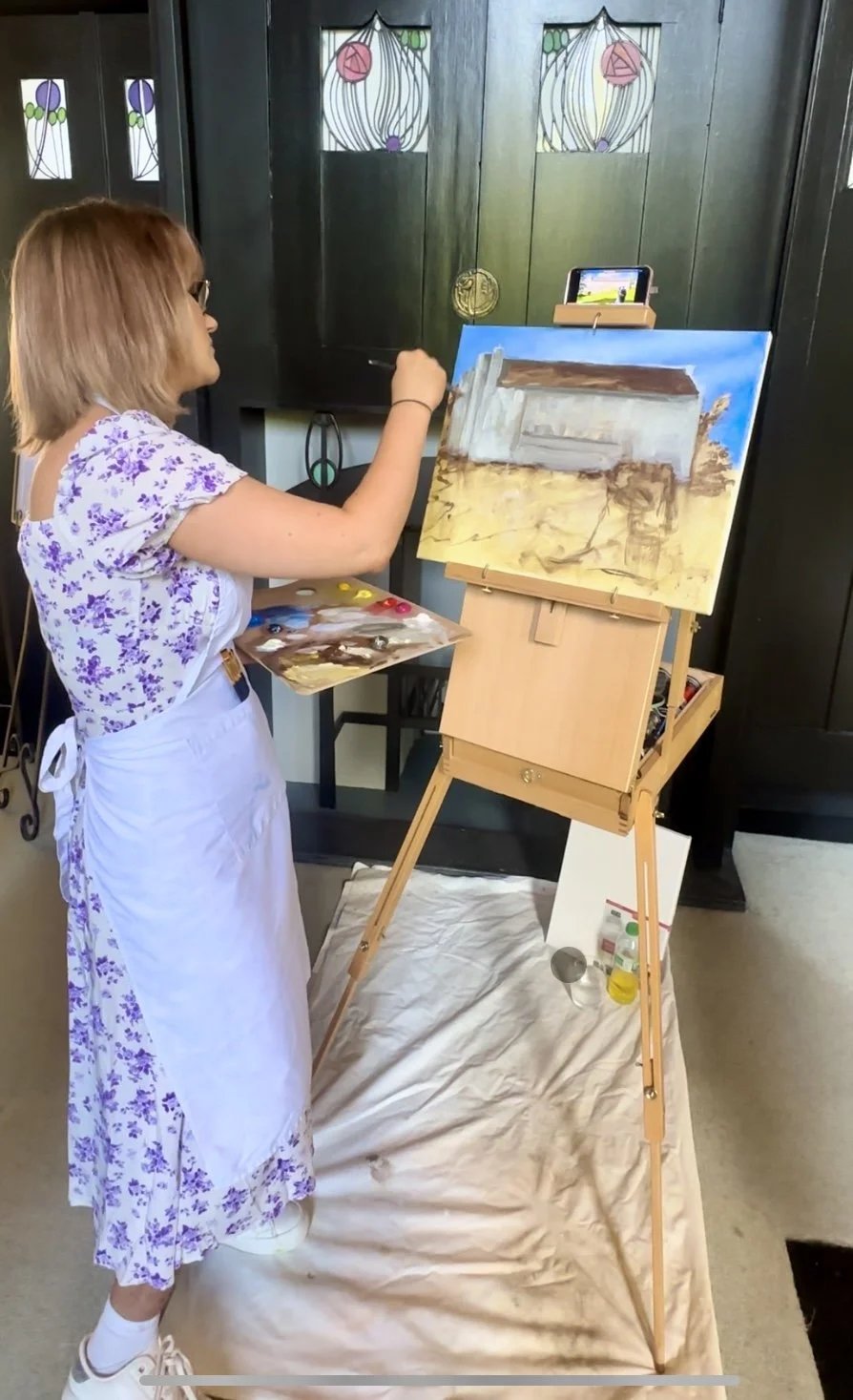 A woman with shoulder-length light brown hair wearing glasses, a white apron, and a white dress with purple flowers, is painting on a canvas set on an easel. The background features dark cabinets with stained glass panels. The woman is holding a paint palette and brush, working on a landscape painting of a barn with a blue sky.
