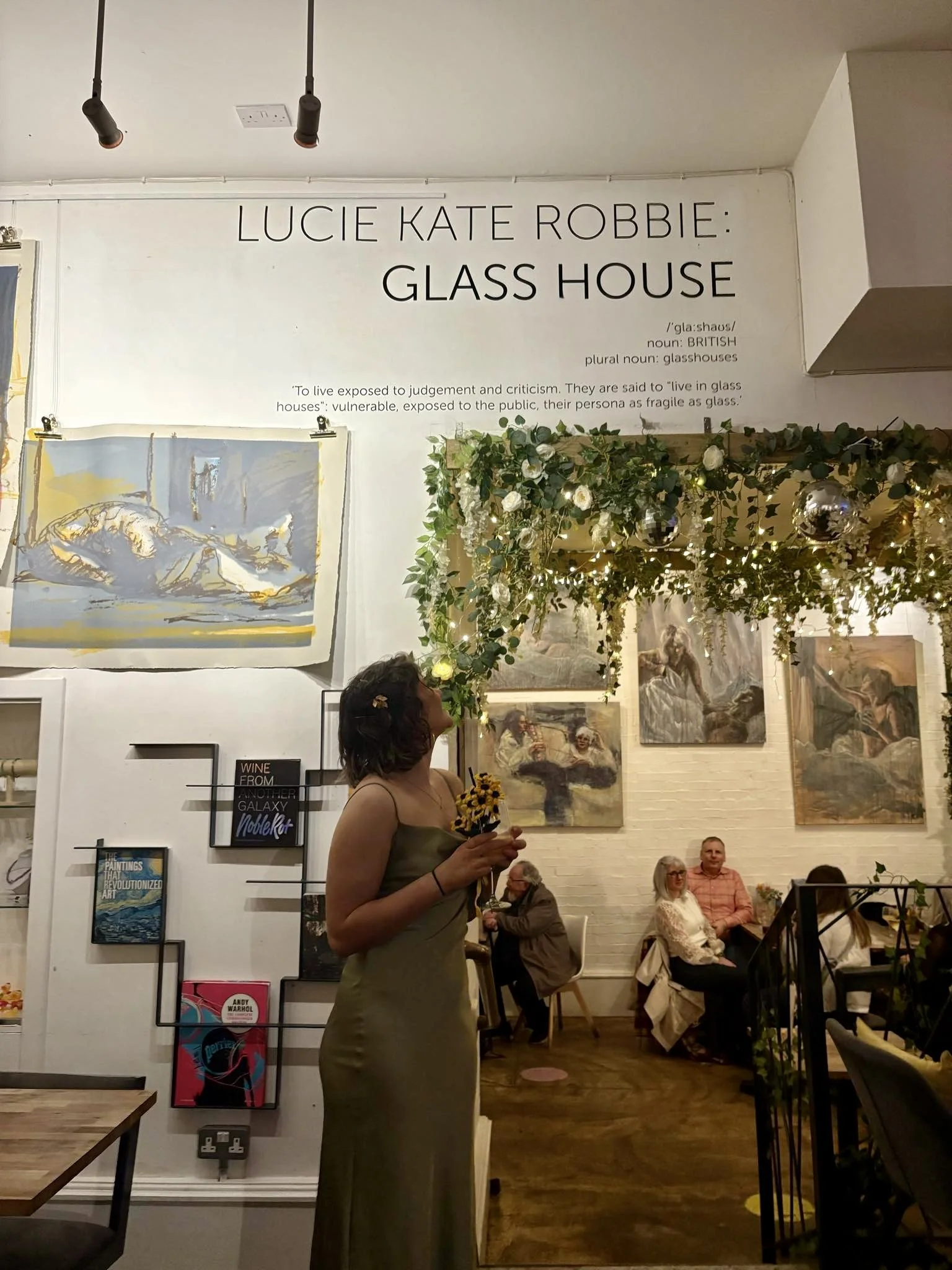 Inside a gallery or cafe with white walls decorated with paintings. A woman in a green dress holding a bunch of yellow flowers is in the foreground. Several people are seated at tables in the background, and a sign on the wall reads "Lucie Kate Robbie: Glass House".