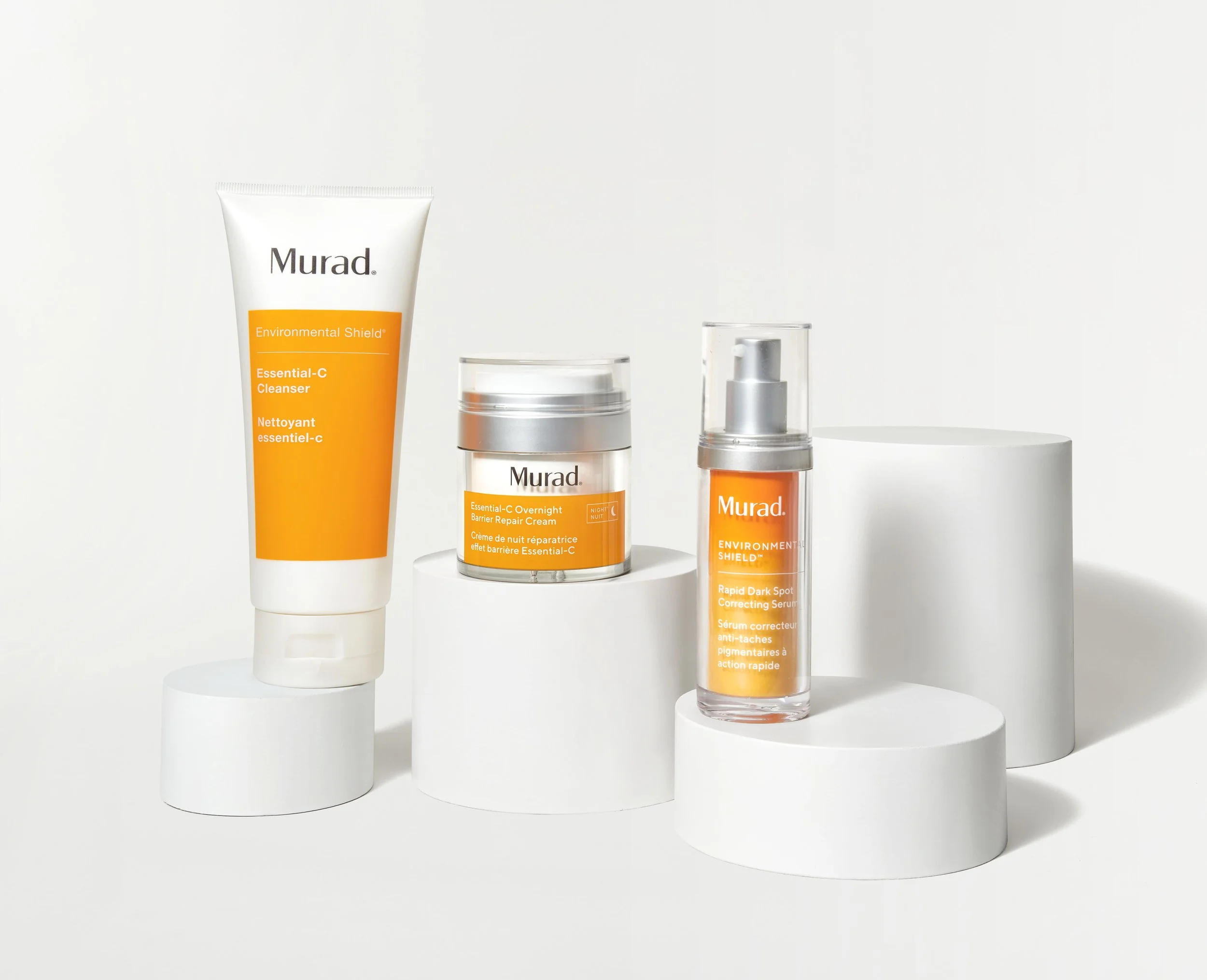 Three skincare products from Murad with orange labels on white cylindrical platforms against a white background.