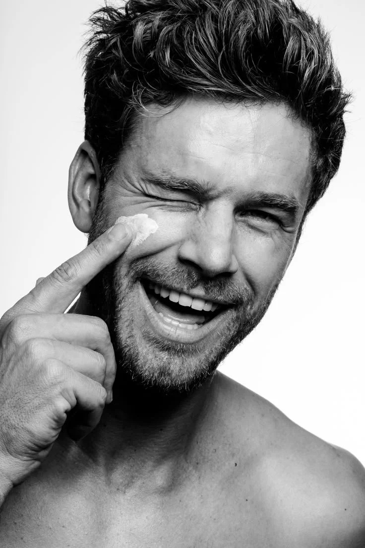 A smiling shirtless man with dark, wavy hair and a beard, applying cream to his cheek with his finger.