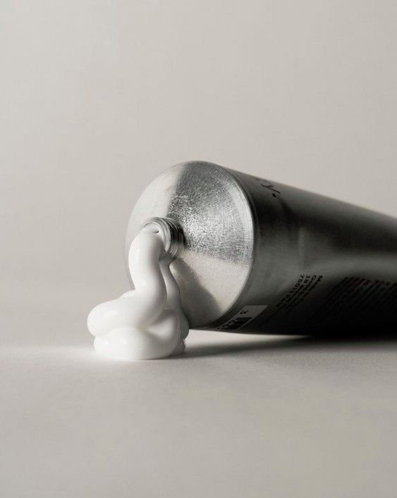 An aluminum tube with white foam spilling out from the opening, lying on its side on a flat surface against a plain background.