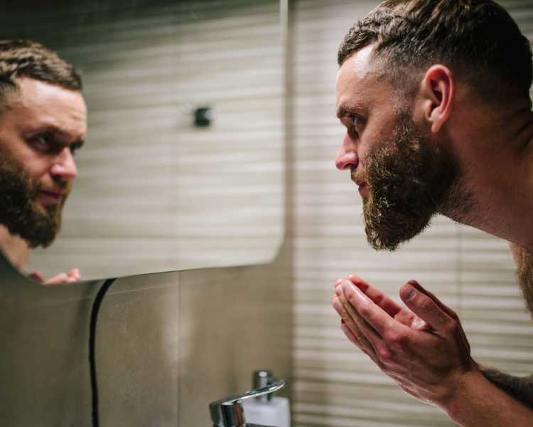 A man with a beard looking into a bathroom mirror, washing his face, with a beige tiled wall in the background.