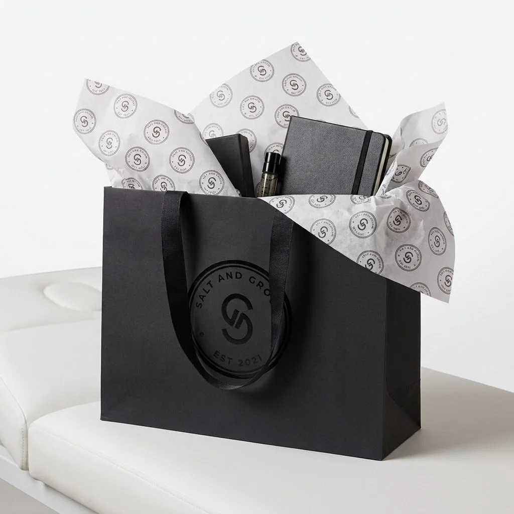 Black gift bag with Salt and Grit logo, containing wrapped tissue paper and three black notebooks or planners with elastic bands, along with a small spray bottle.