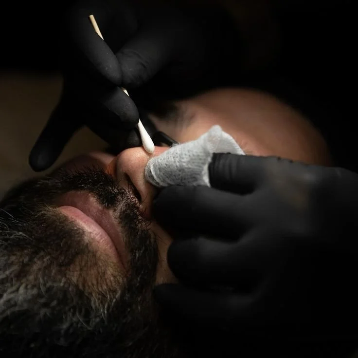 A person getting a nose piercing while lying down, with a gloved technician cleaning the nose with a cotton swab.