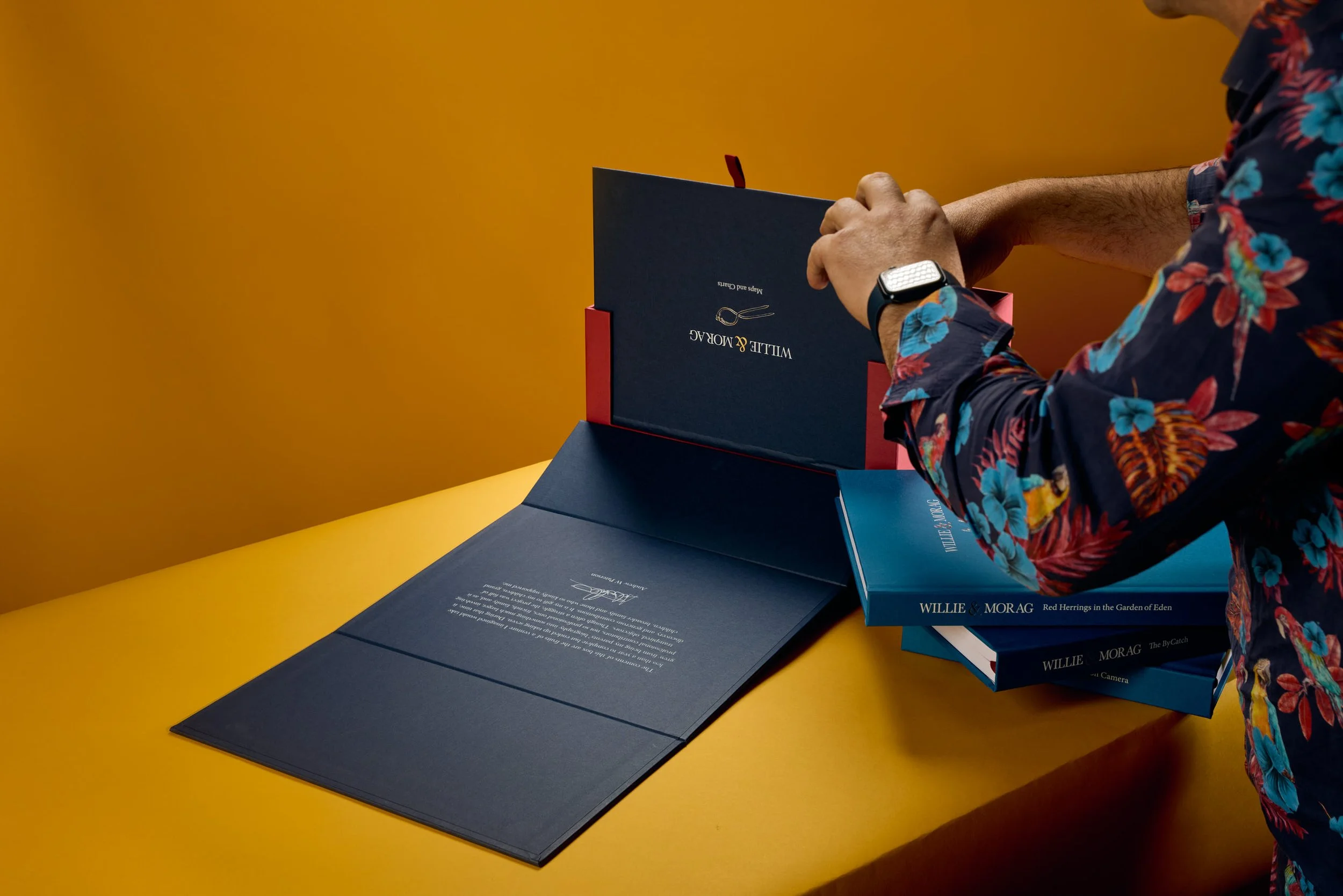 Person in a floral shirt opening a navy blue box labeled 'Willie & Morag' against a yellow background, with opened books and document on the table.