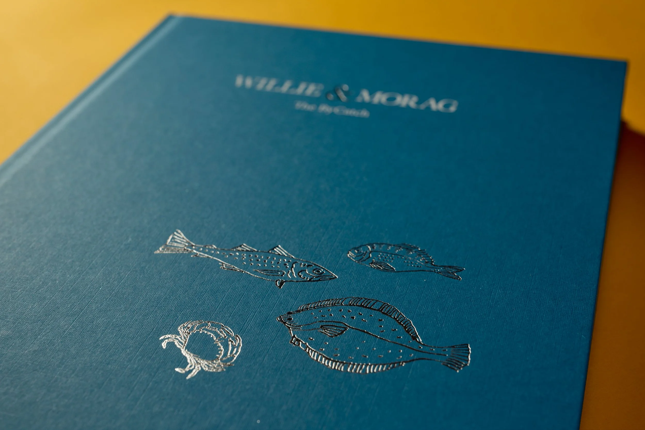 A blue notebook or journal with silver embossed fish illustrations and the text 'FISHERMAN'S LOGBOOK' in silver.