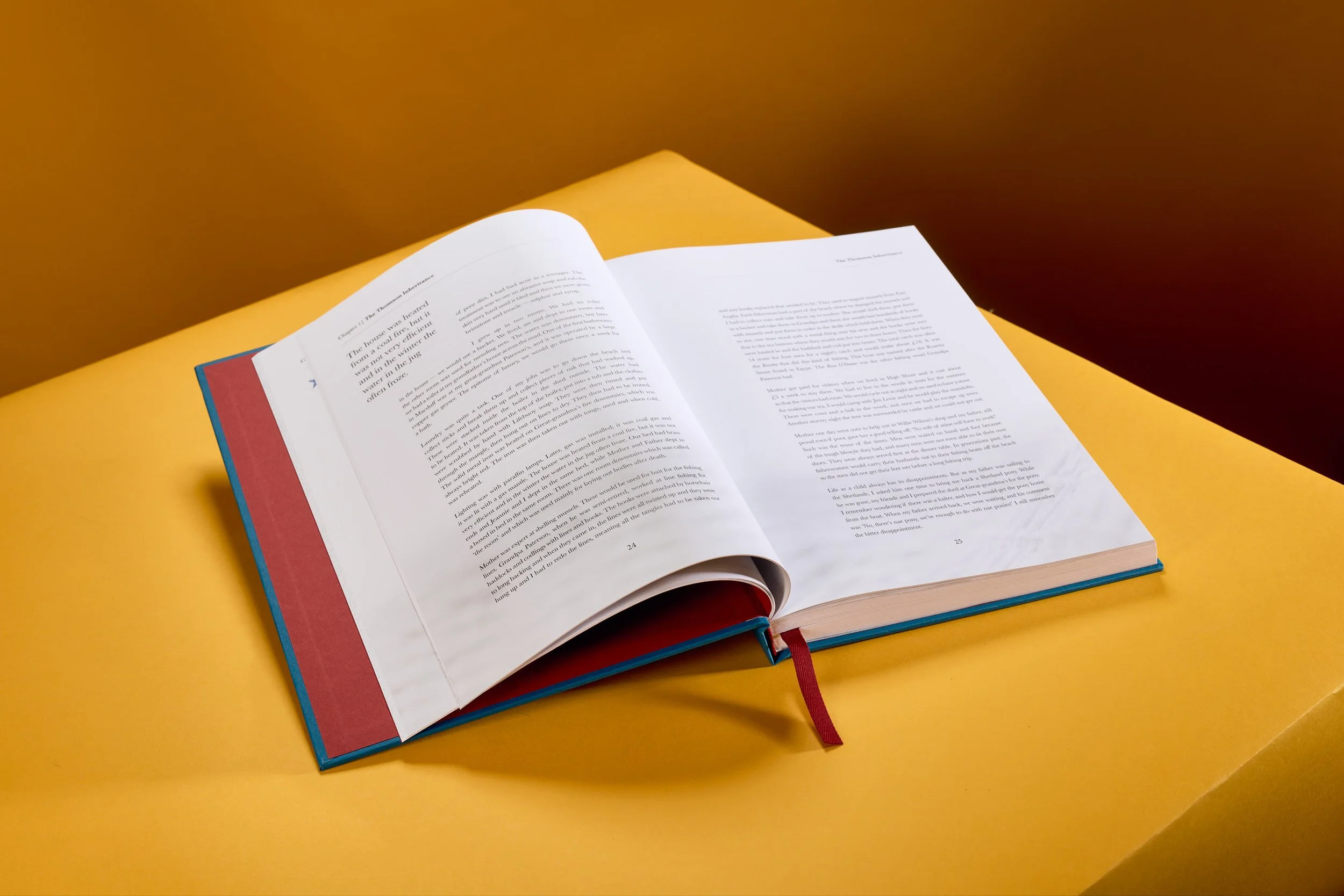 An open hardcover book with white pages resting on a yellow table. The book has a red ribbon bookmark, and a blue cover with a red and blue edge is visible underneath.