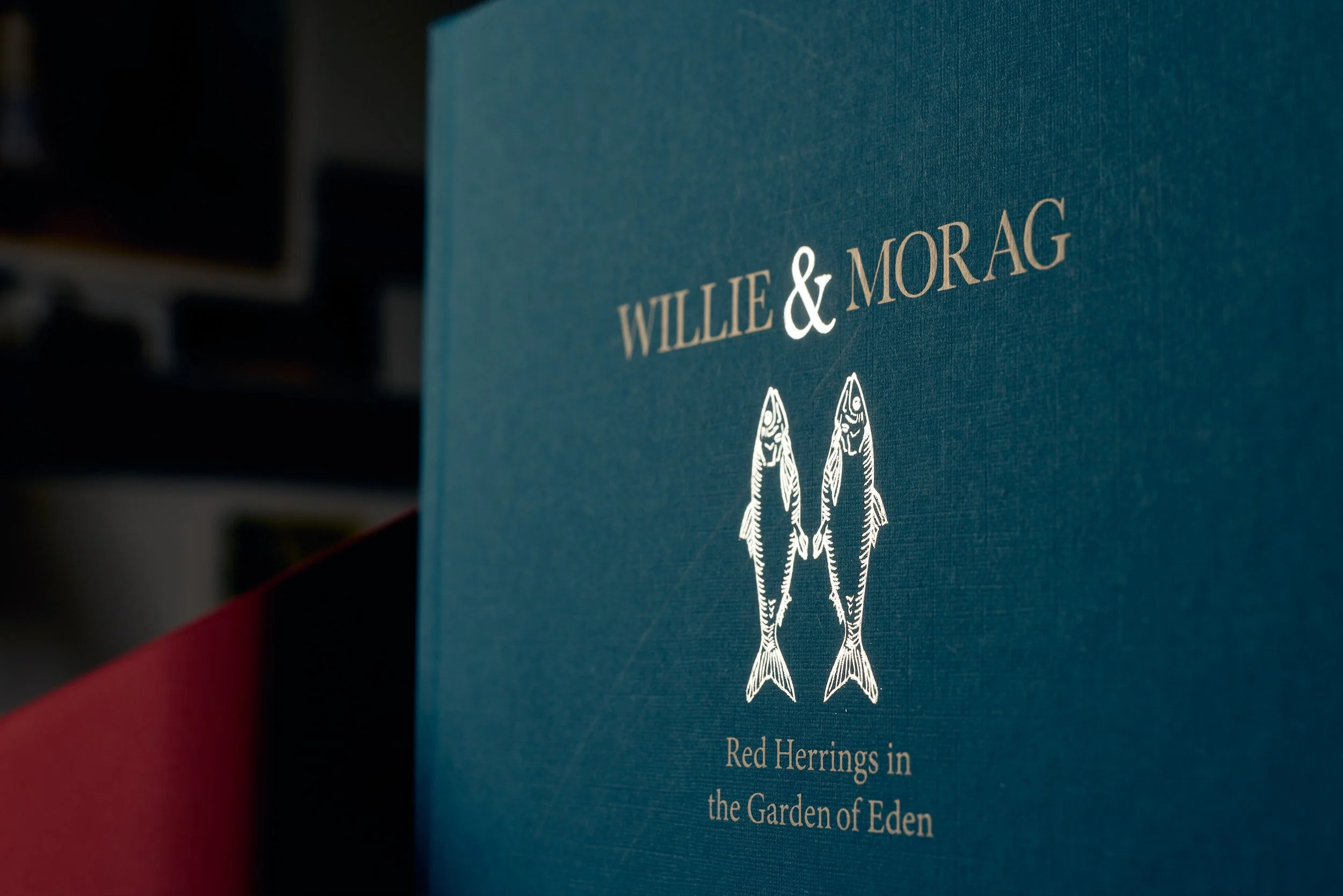 Close-up of a book cover titled 'Willie & Morag' with illustrations of two fish and the subtitle 'Red Herrings in the Garden of Eden'.