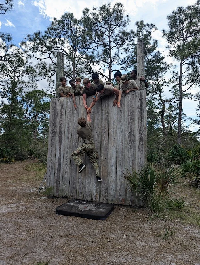 A person in camouflage pants and a T-shirt is climbing a wooden wall while others in military uniform are cheering from the top.