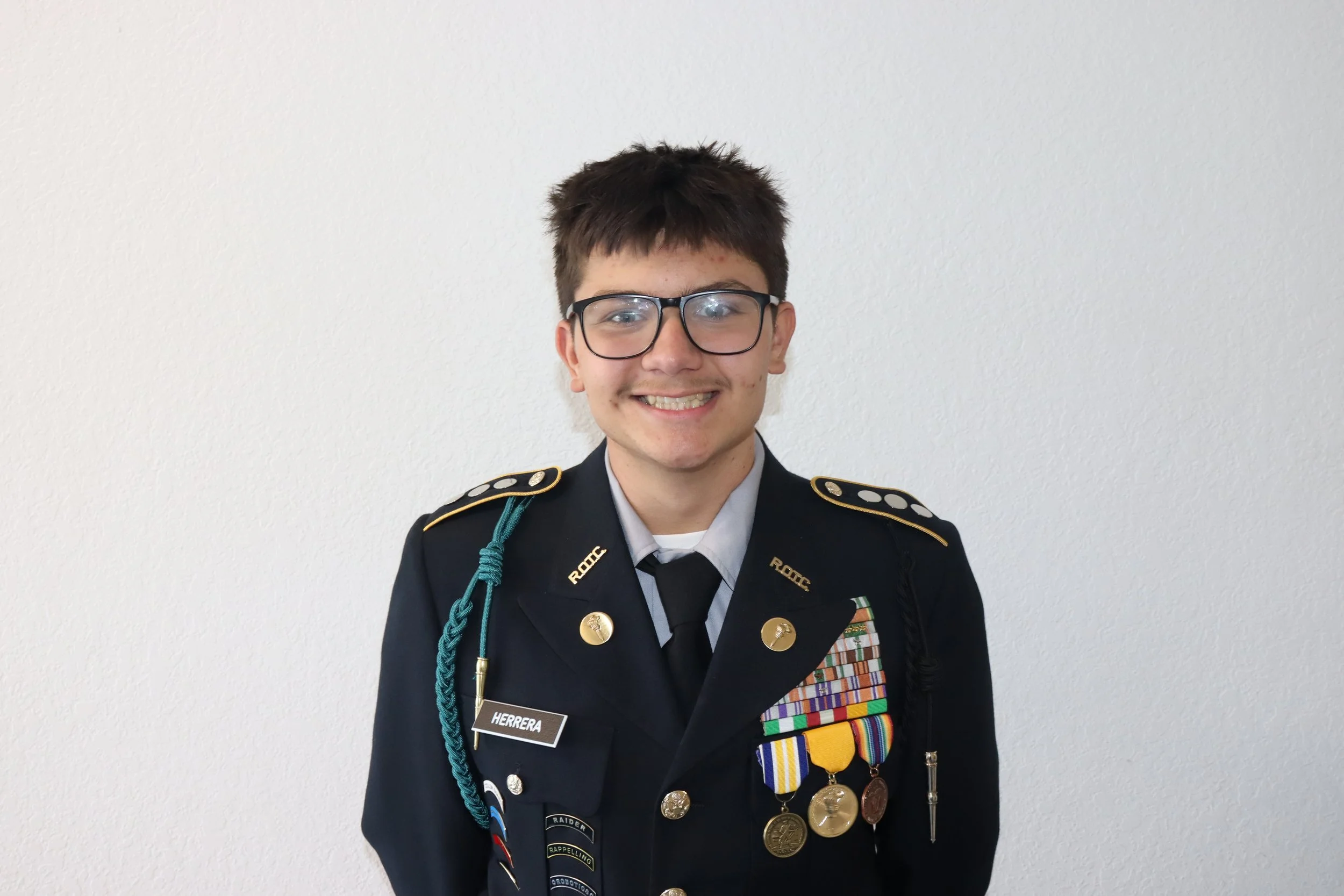 Young boy in military uniform with medals and ribbons, smiling, standing against a plain white wall.